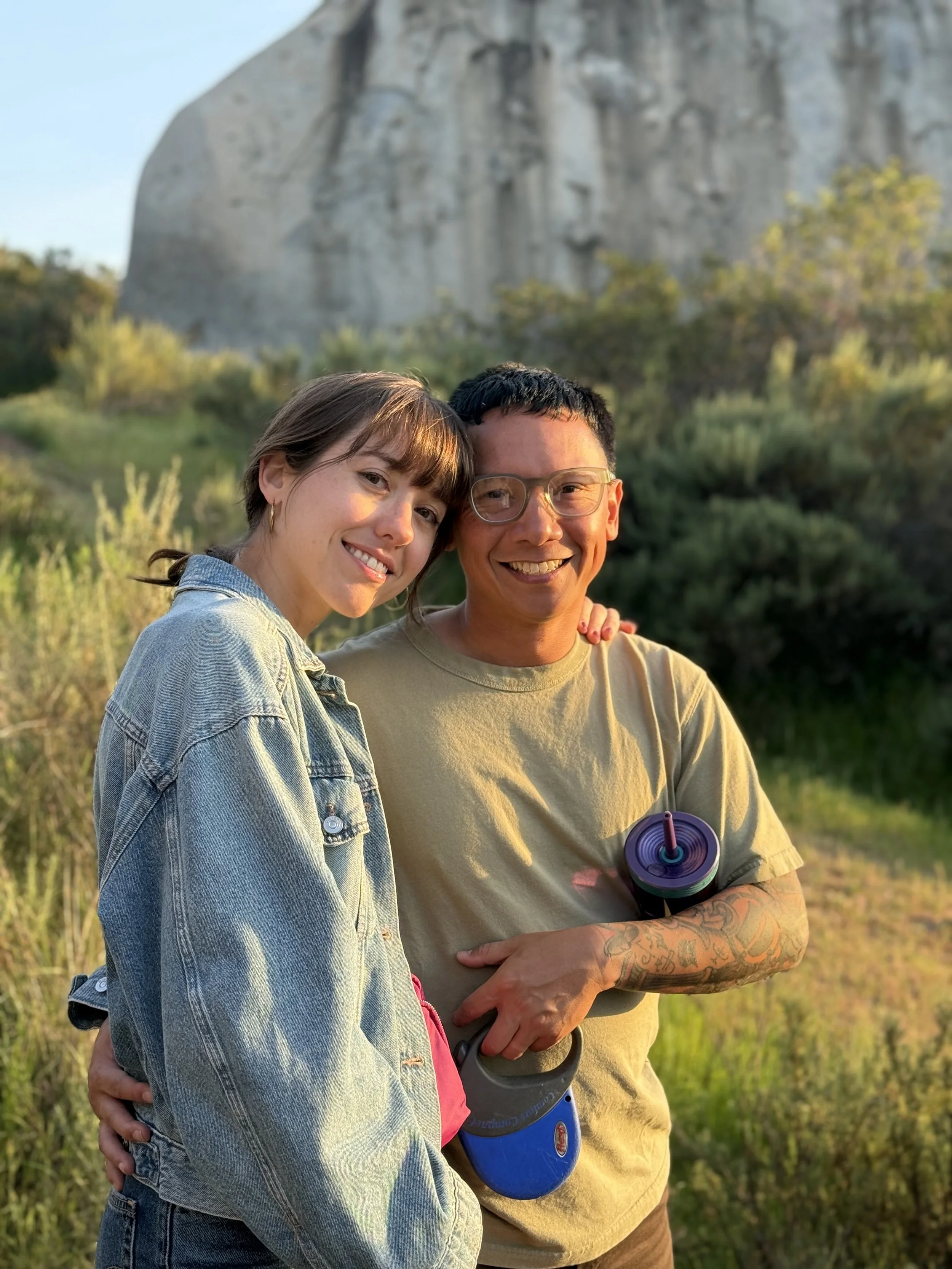 Two people, a woman and a man, smiling and standing close together outdoors in a green, natural setting with trees and a large rock formation in the background. The woman has shoulder-length brown hair, is wearing a denim jacket and gold hoop earrings. The man has short black hair, glasses, tattoos on his arm, and holds a blue and black water bottle.