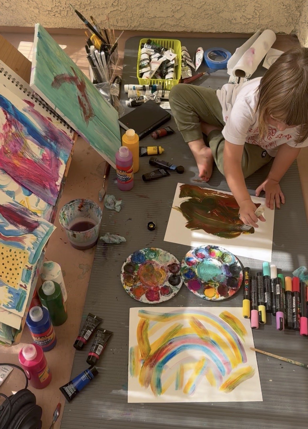 Child creating abstract paintings on paper with various paint colors and brushes on the floor.