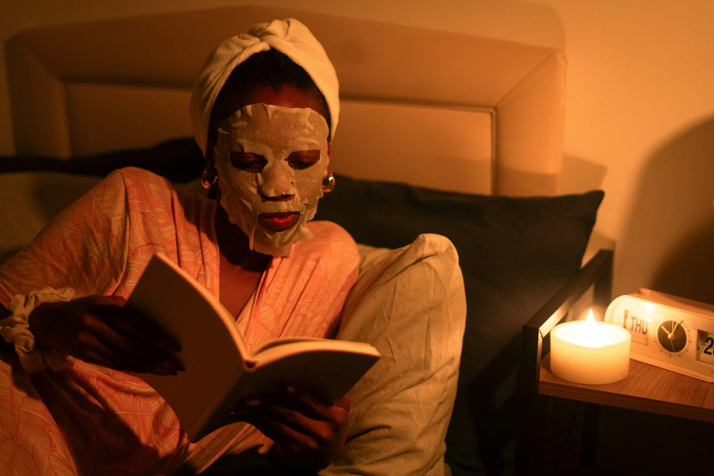 A person with a facial sheet mask, a towel wrapped around their head, wearing earrings, reading a book in a dimly lit room with candles on the side.