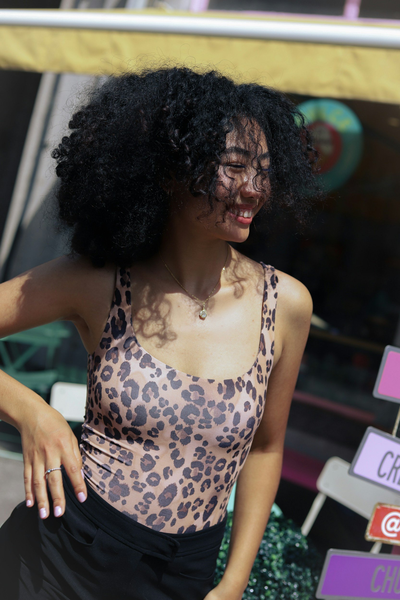 A woman with curly black hair smiling, wearing a leopard print tank top and black pants, in front of a display with signs.