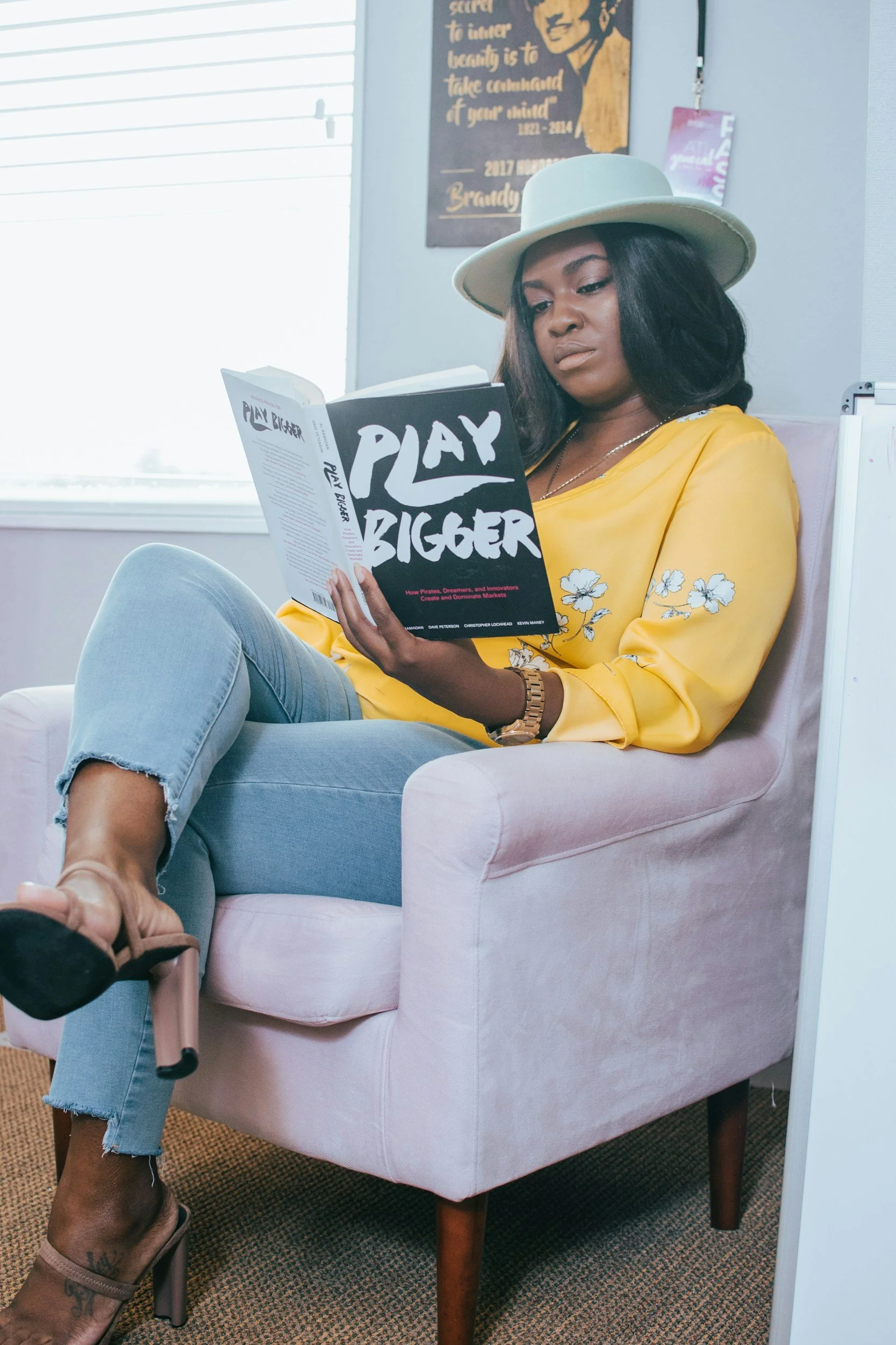 Woman with dark hair, wearing a white hat and yellow floral top, sitting in a pink armchair, reading a book titled 'Play Bigger' in an office or lounge with a window and wall decorations.