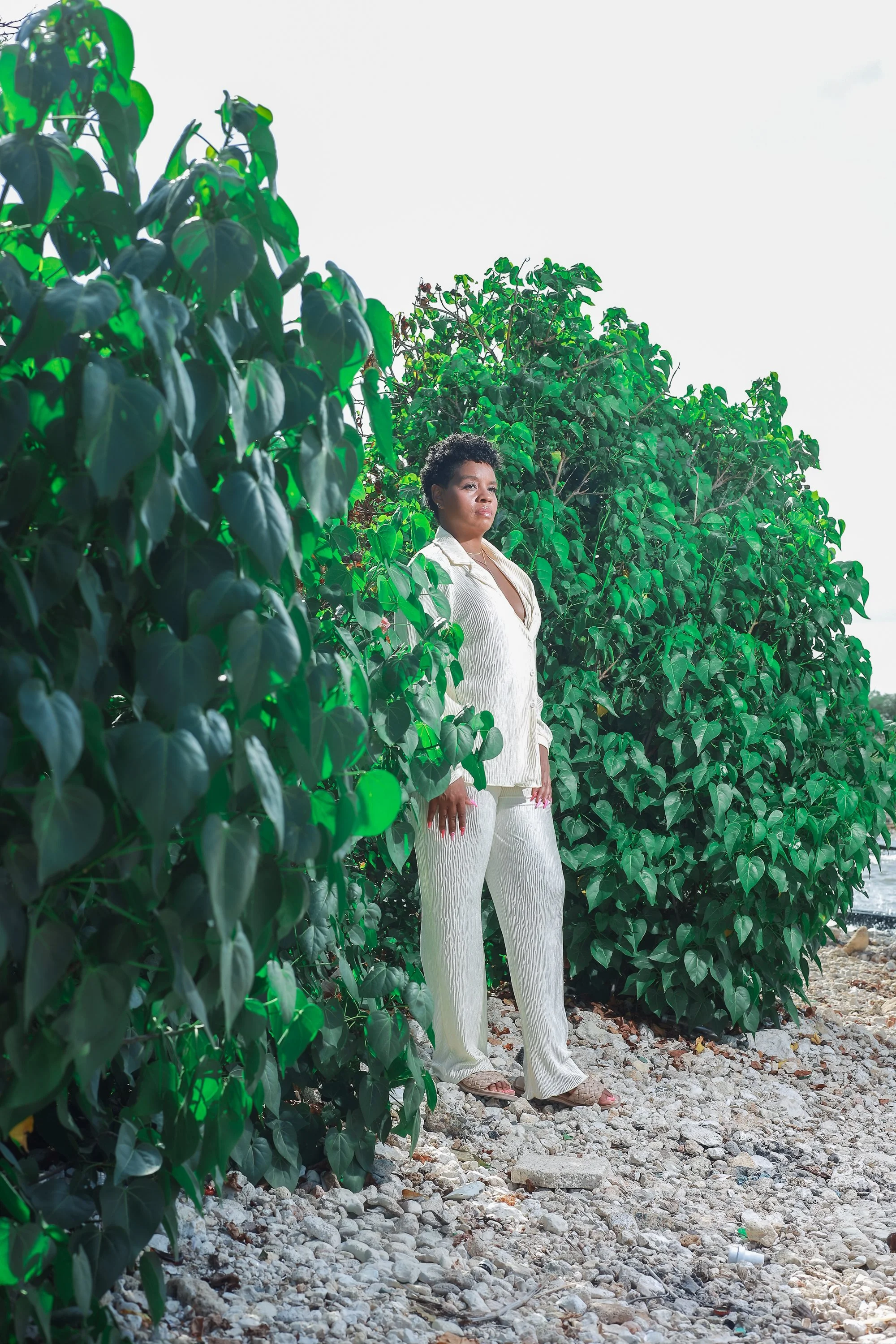 A woman standing among green bushes on rocky terrain under a cloudy sky.