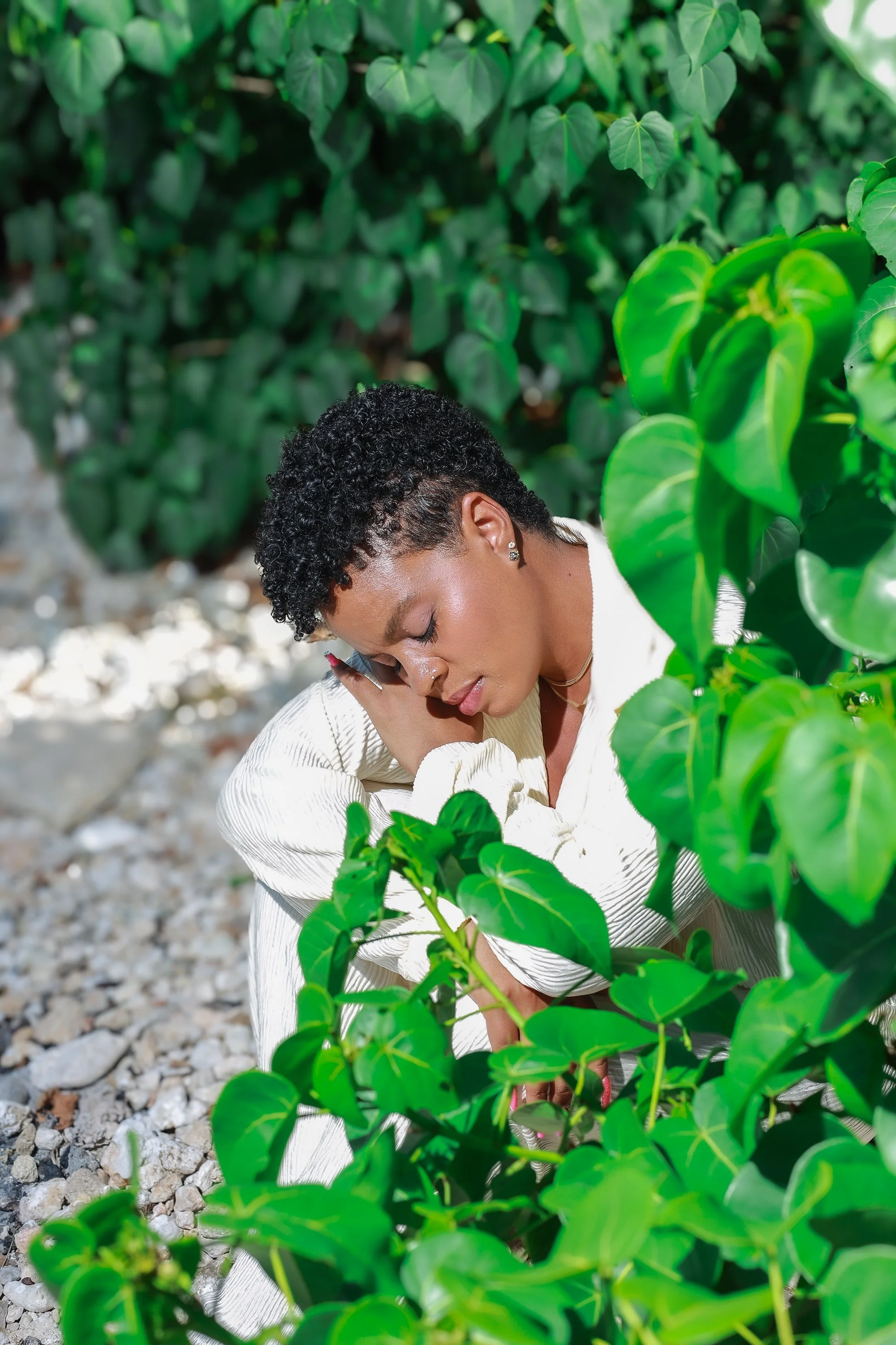 A woman with short curly black hair and earrings, sitting outdoors on a rocky surface, surrounded by green leafy plants, with her eyes closed and her head resting against her hand.