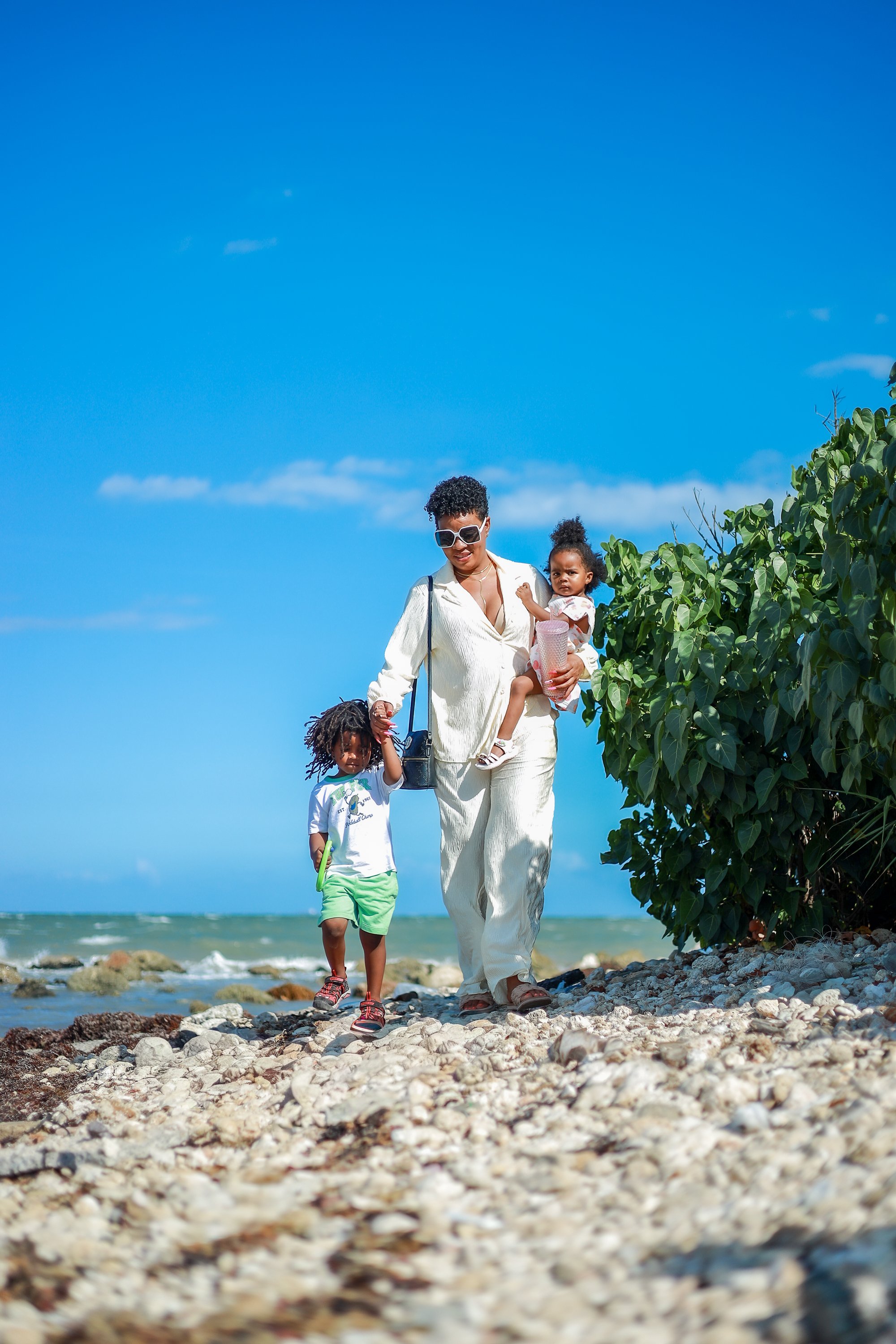 A woman wearing white sunglasses and white clothing is walking along a rocky beach with two children. She is carrying a young girl in her right arm, and the girl has curly hair and is holding a pink water bottle. The other child, a boy with curly hair, is walking beside her and is dressed in a white t-shirt and green shorts. There is green foliage to the right, and the ocean and a clear blue sky are in the background.