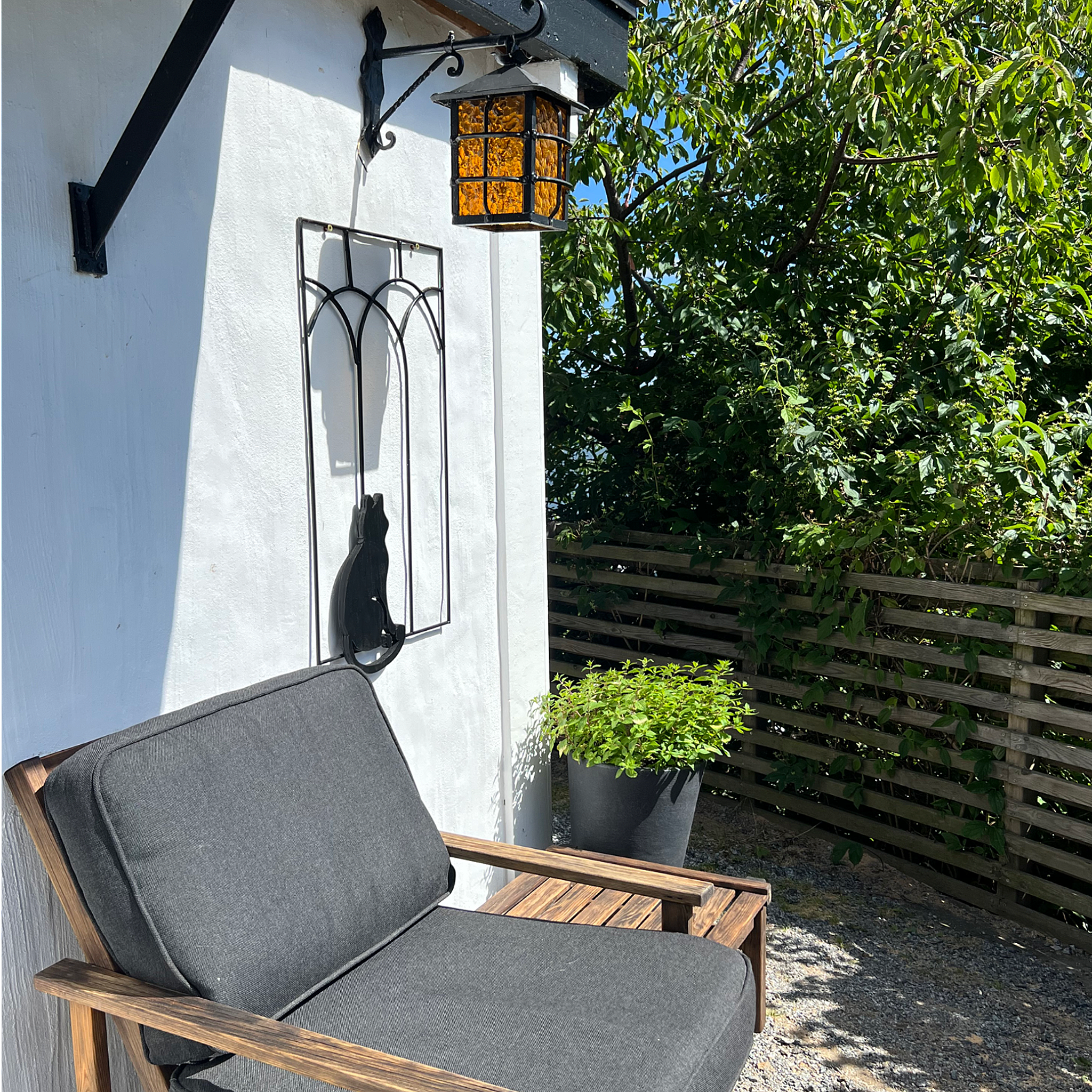 Utomhusplats med en trästol med mörkgrå sittdyna, ett svart metallstativ för växter, en facklampsliknande lykta med orange glas, ett träbord, gröna träd och ett trädstaket i bakgrunden.