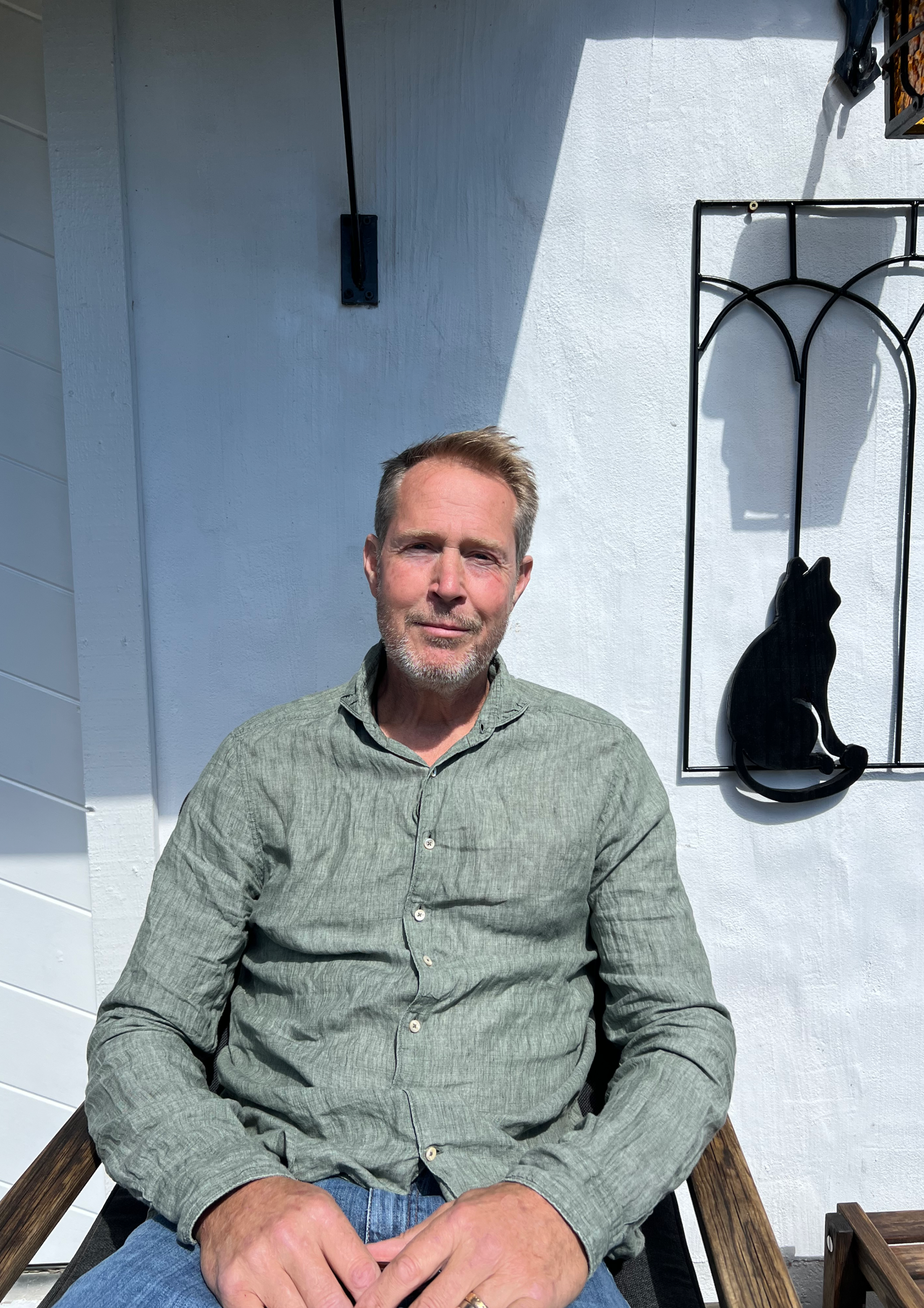 Man sitter på en stol utomhus framför en vit vägg med skuggor och metalldekoration.