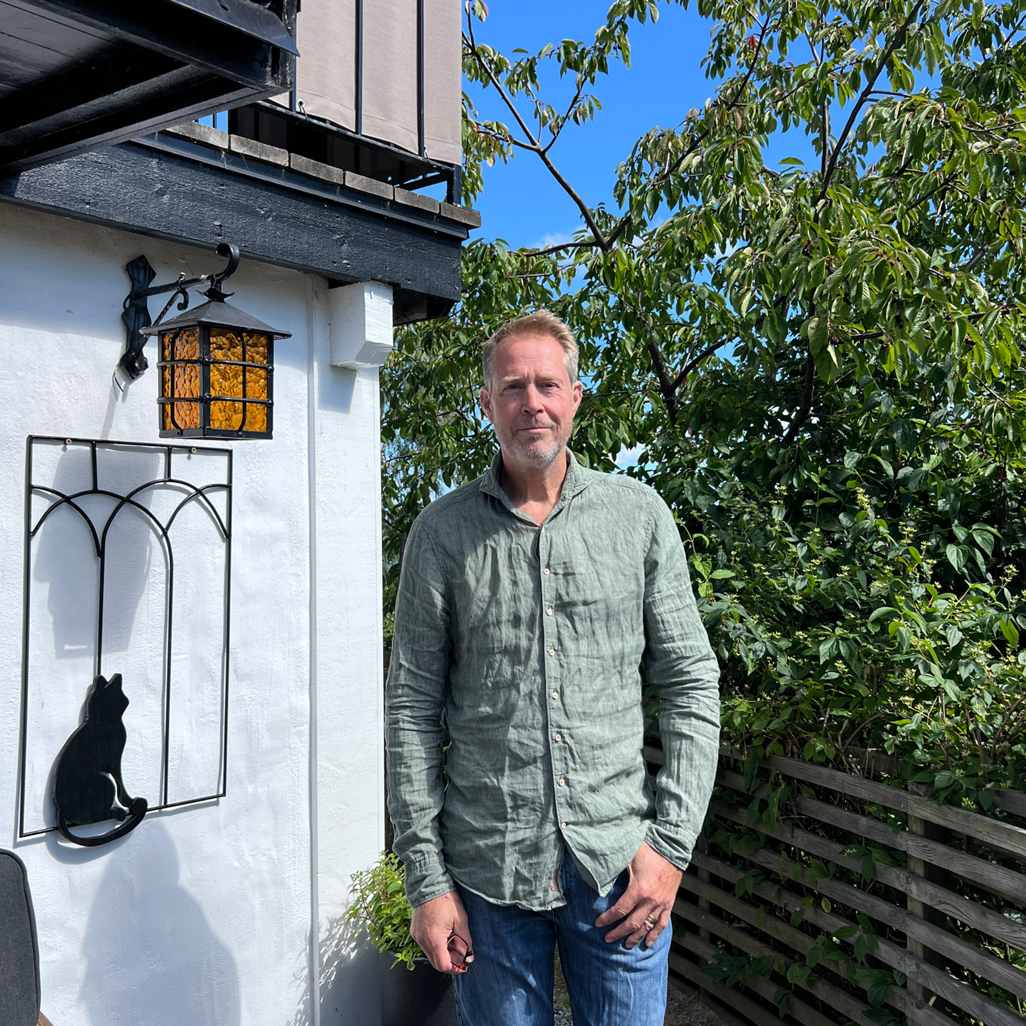 En man står utomhus framför ett trästaket, med ett träd med gröna blad i bakgrunden, och ett svart väggfäste med ett oljelampa. Mannen tittar mot kameran och är klädd i grå skjorta.