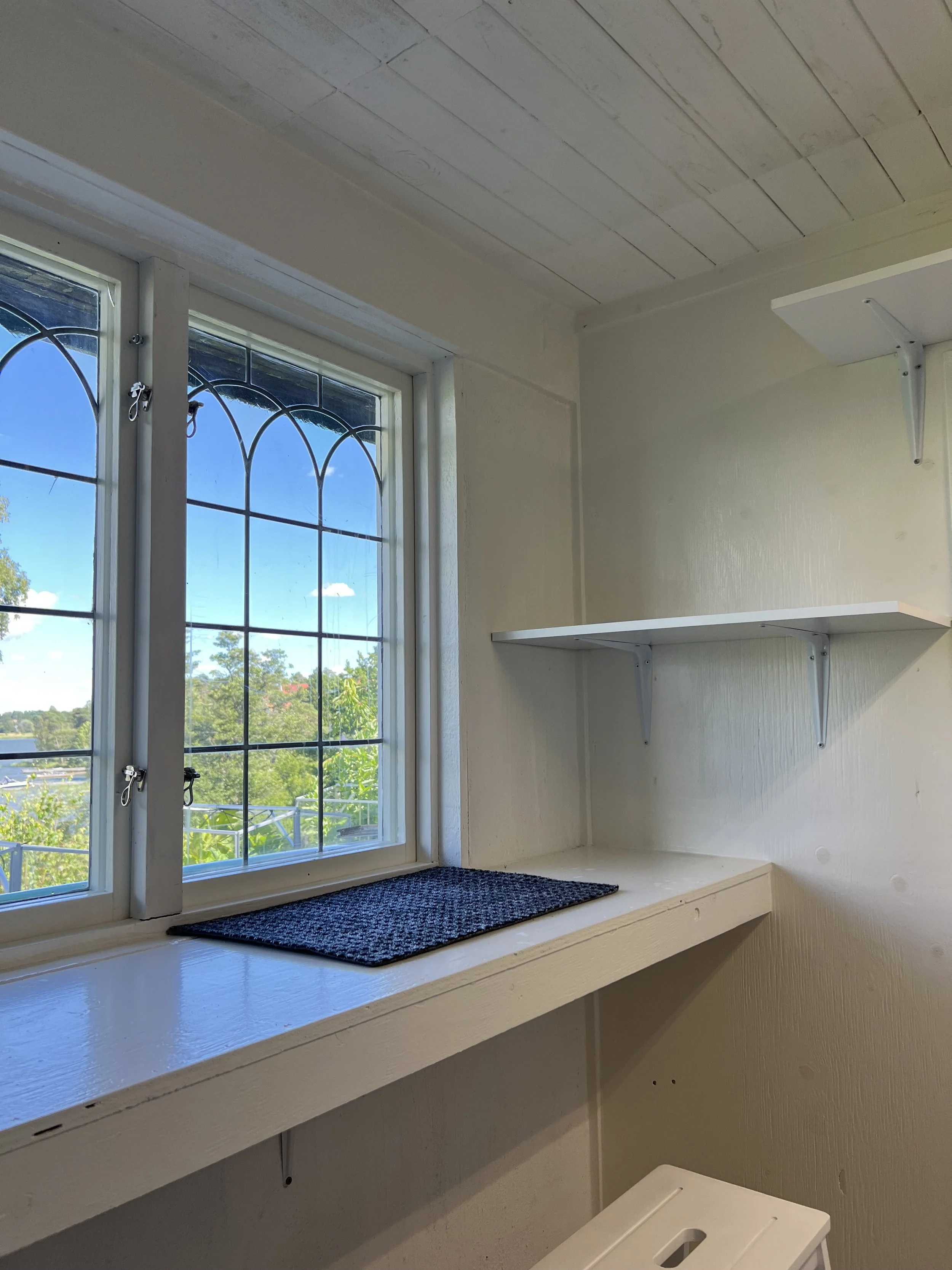 Ett fönster med utsikt över gröna träd och en blå himmel, samt en vit hylla och en svart matta på fönsterbrädan.