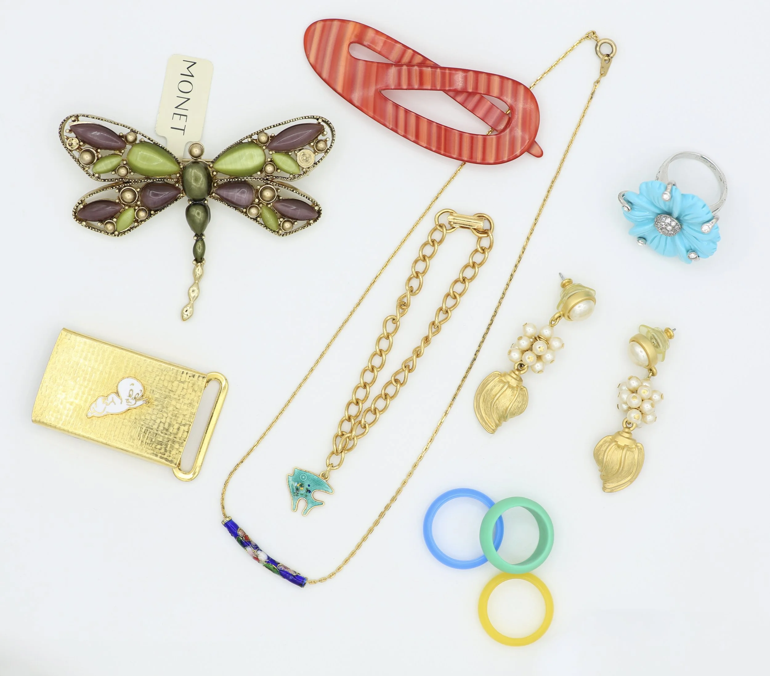 Collection of jewelry including a colorful butterfly pendant with purple and green stones, a coral-colored hair clip, gold chains, a flower-shaped ring with blue petals and a clear center, pearl earrings, a watch with a gold band, a keychain with a blue dolphin, and colorful plastic rings on a white background.