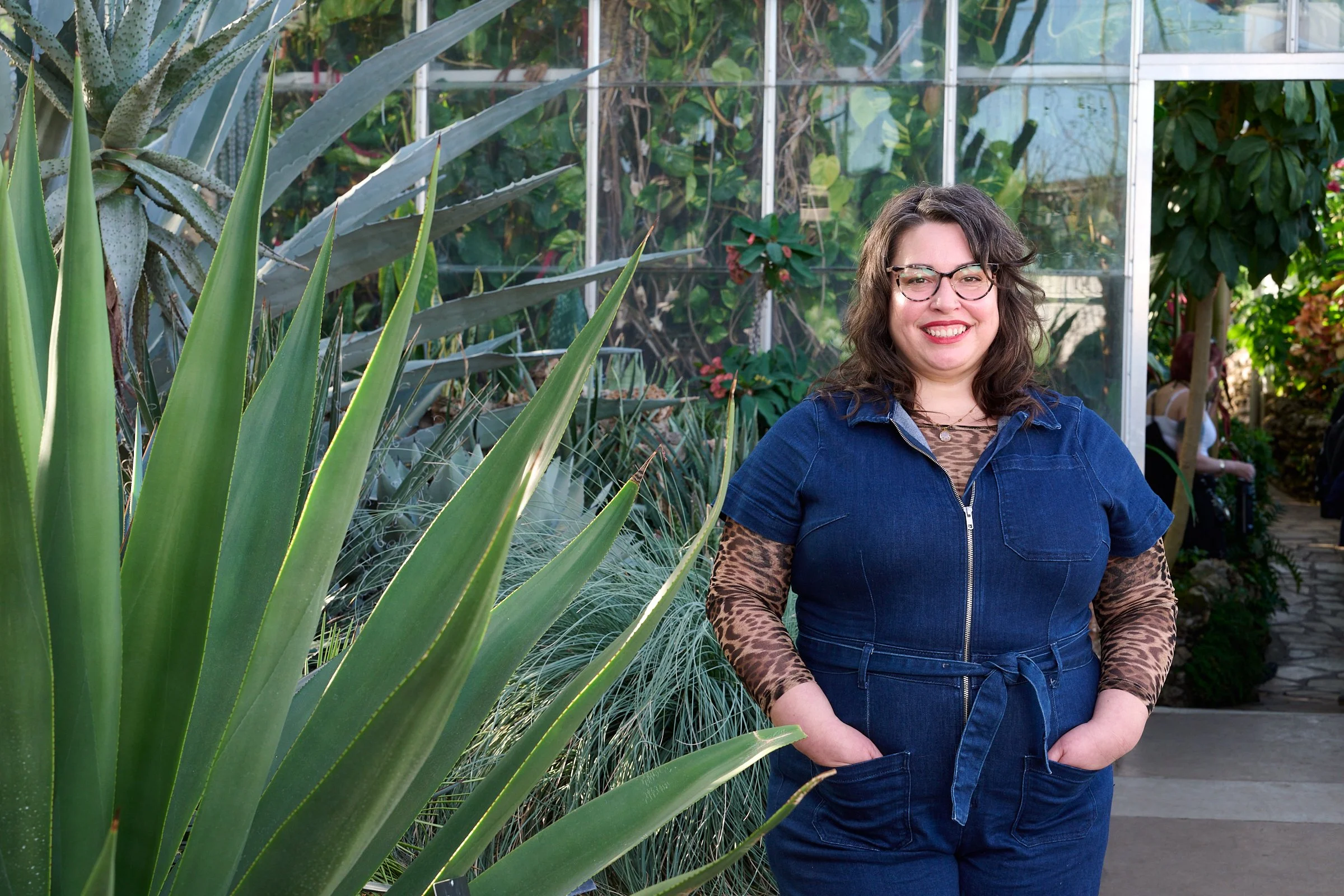 A woman with glasses and wavy brown hair smiling at the camera in a greenhouse surrounded by large succulents and green plants.