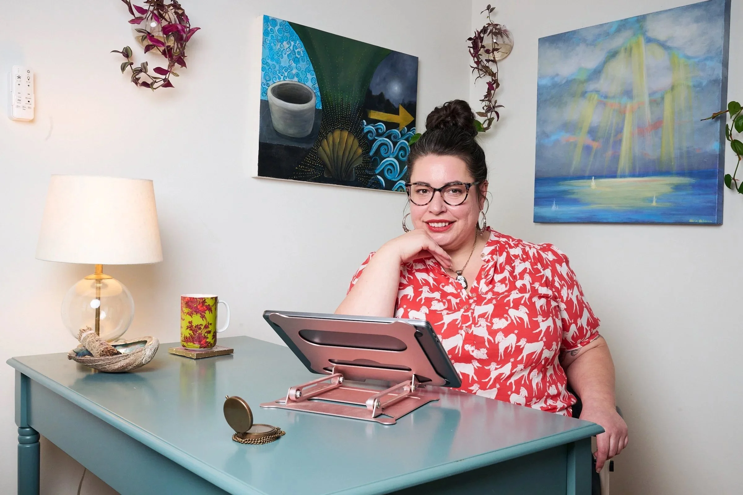 A woman with glasses and earrings, wearing a red blouse with white horse silhouettes, sitting at a light blue desk with a tablet. The desk has a lamp, a decorative bowl, a colorful mug, and a gold clip. Behind her are paintings and potted plants, with a wall-mounted remote control visible on the left.