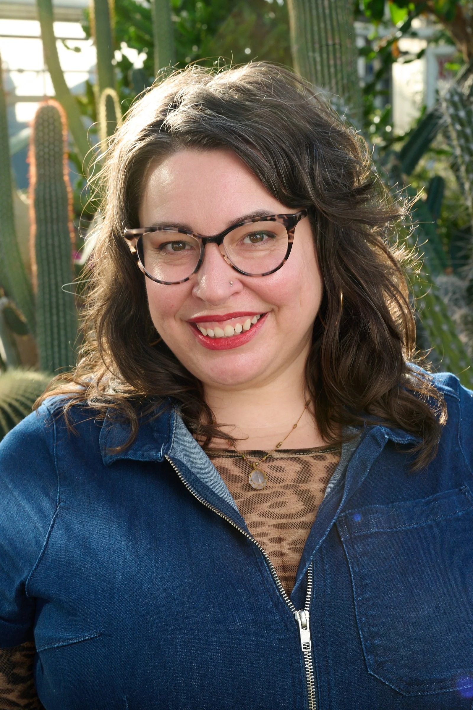 Close-up of a smiling woman with curly brown hair, wearing glasses, a nose piercing, a denim jacket, and layered necklaces, standing outdoors with cactus plants in the background.