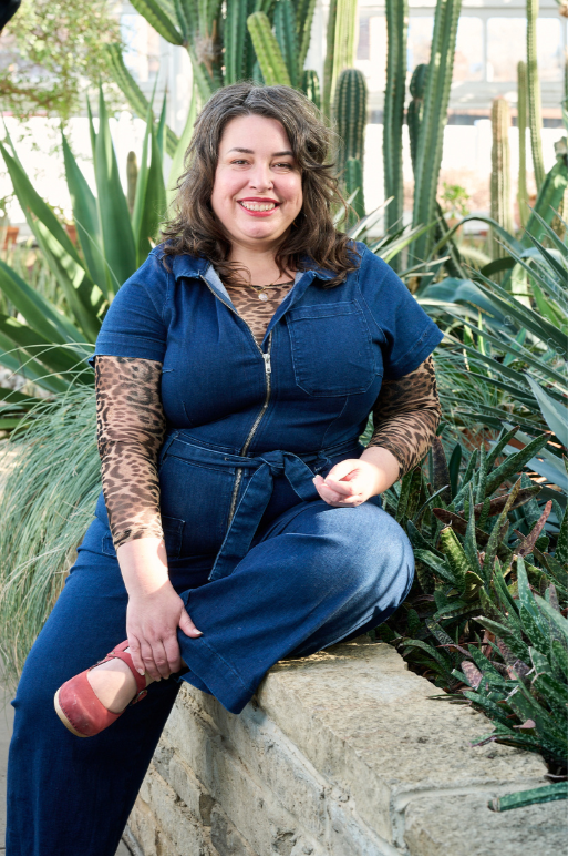 A woman with shoulder-length wavy brown hair, smiling, wearing a denim jumpsuit over a leopard print long-sleeve shirt, sitting on a stone ledge surrounded by green cacti in a greenhouse.