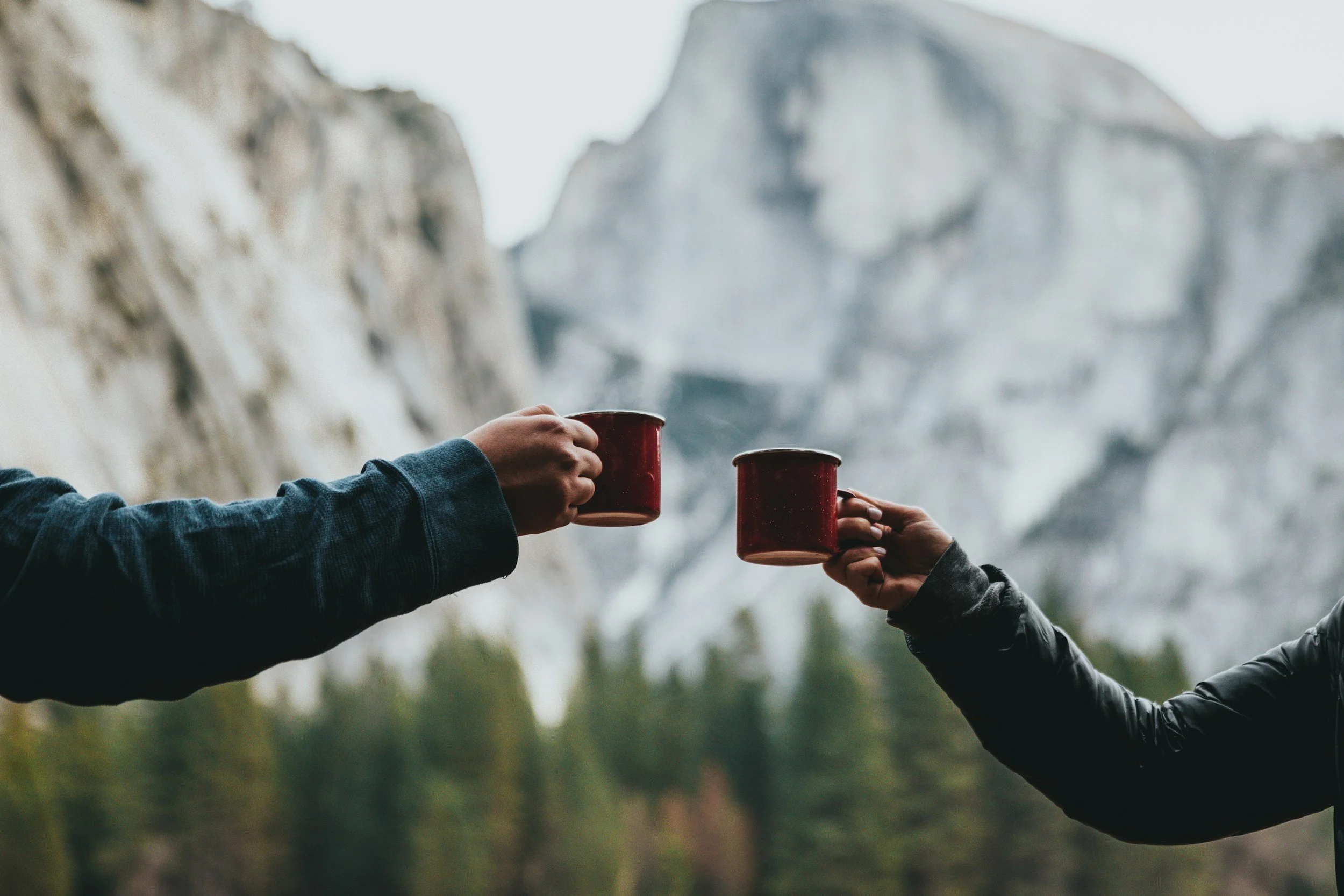 Two people are toasting with camp mugs outdoors with mountains and trees in the background.