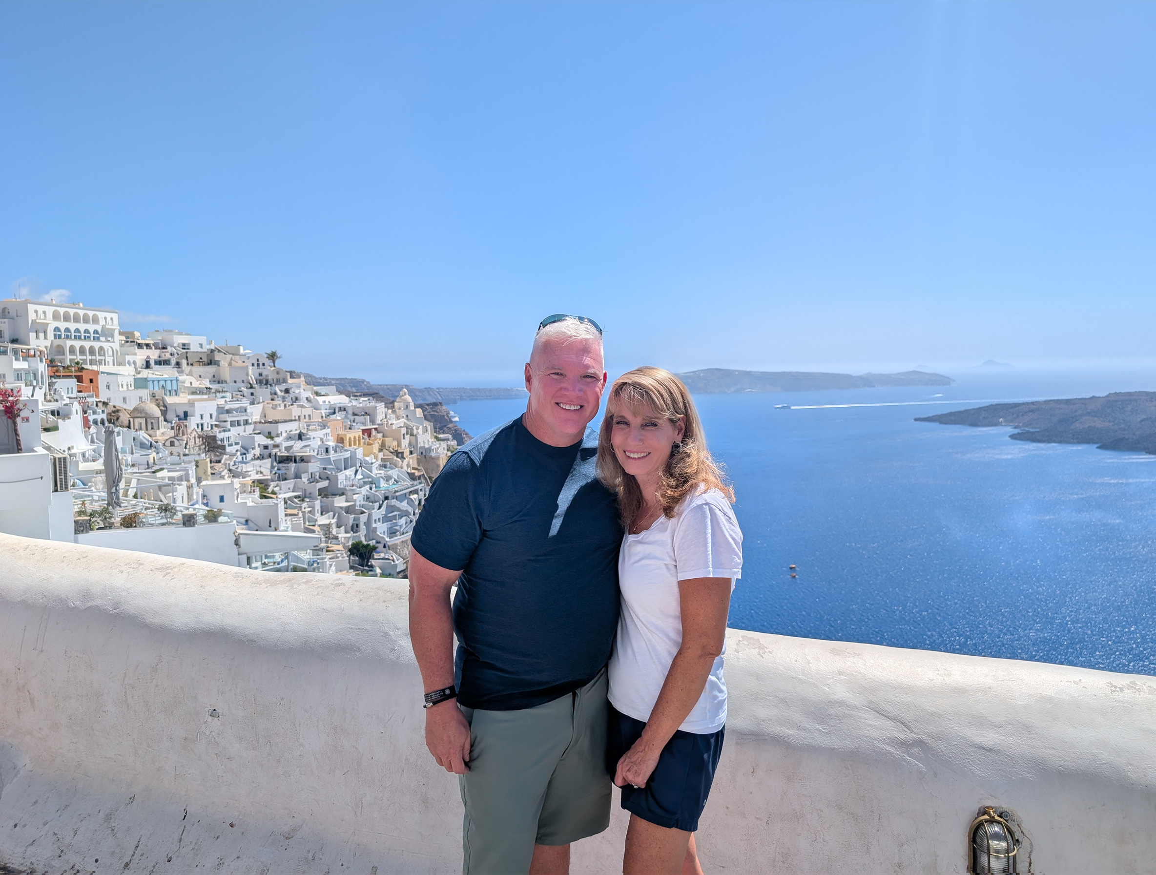 A smiling couple, Founders of Roasted Lantern coffee,  stand close together on a white balcony overlooking the blue Aegean Sea and white buildings of a scenic coastal town, with clear skies above.