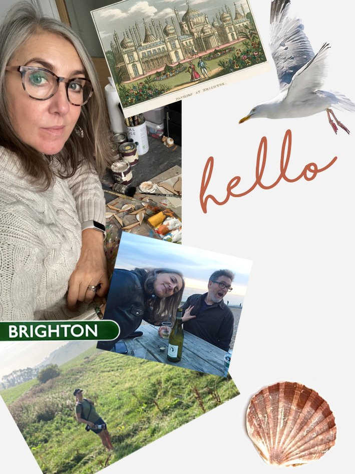 A woman with glasses and a beige sweater takes a selfie in her art studio, surrounded by paintings, art supplies, a seagull illustration with the word "hello," a photo of two people at sunset with wine, a person hiking on a green hillside, and a seashell.