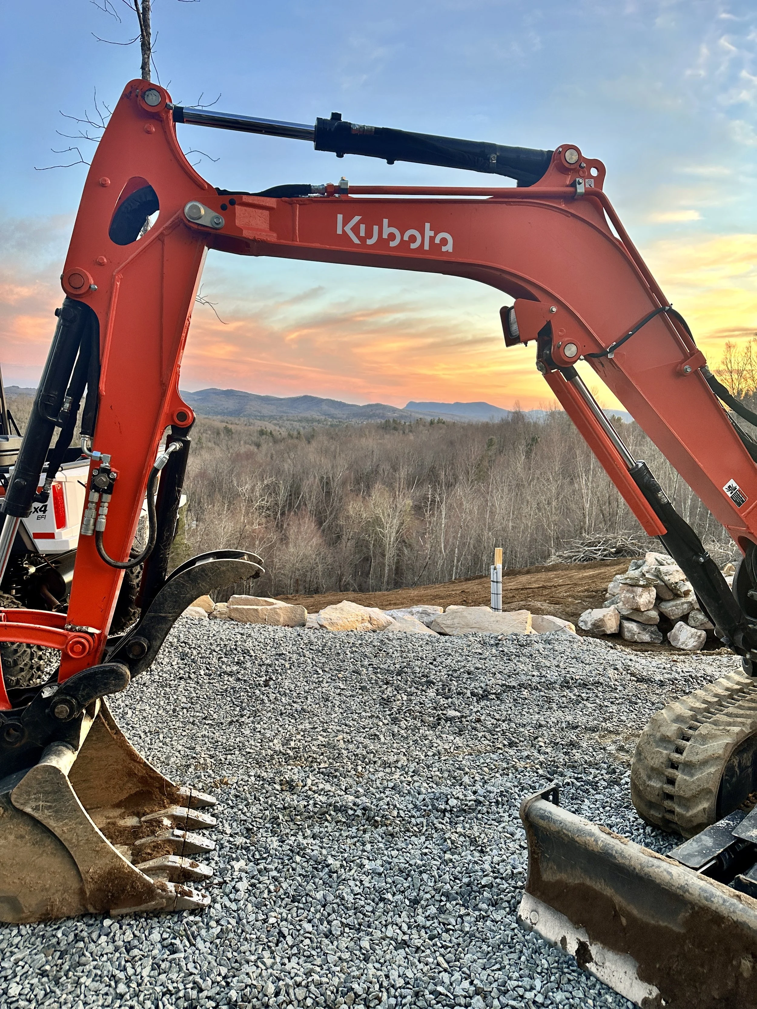 Close-up of orange Kubota excavator arm at a construction site with gravel ground, rocks, and a sunset sky with clouds and mountains in the background.