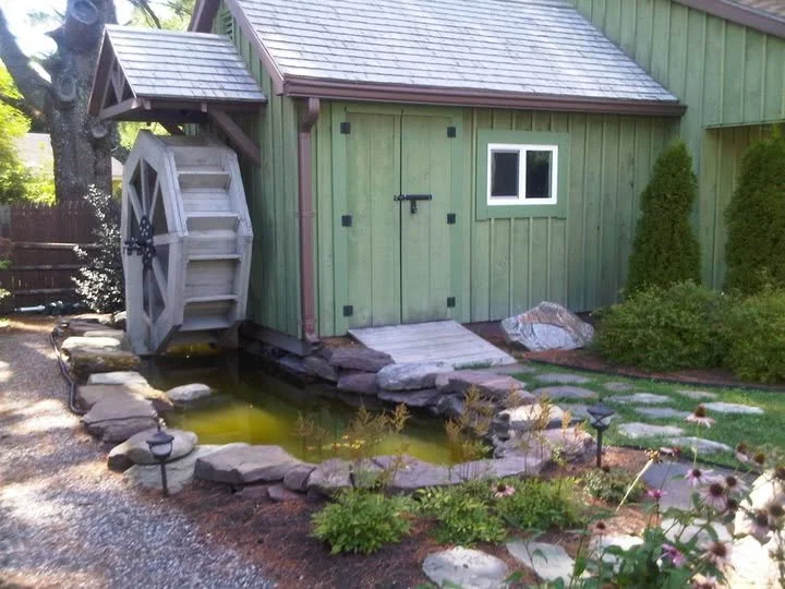 A miniature green watermill with a water wheel beside a small pond, surrounded by plants and a stone pathway.