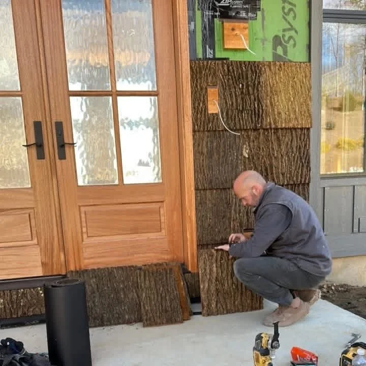 A man crouching outside a house under construction, working on a wooden siding wall near a wood door with glass panels. Tools and construction materials are visible on the ground.