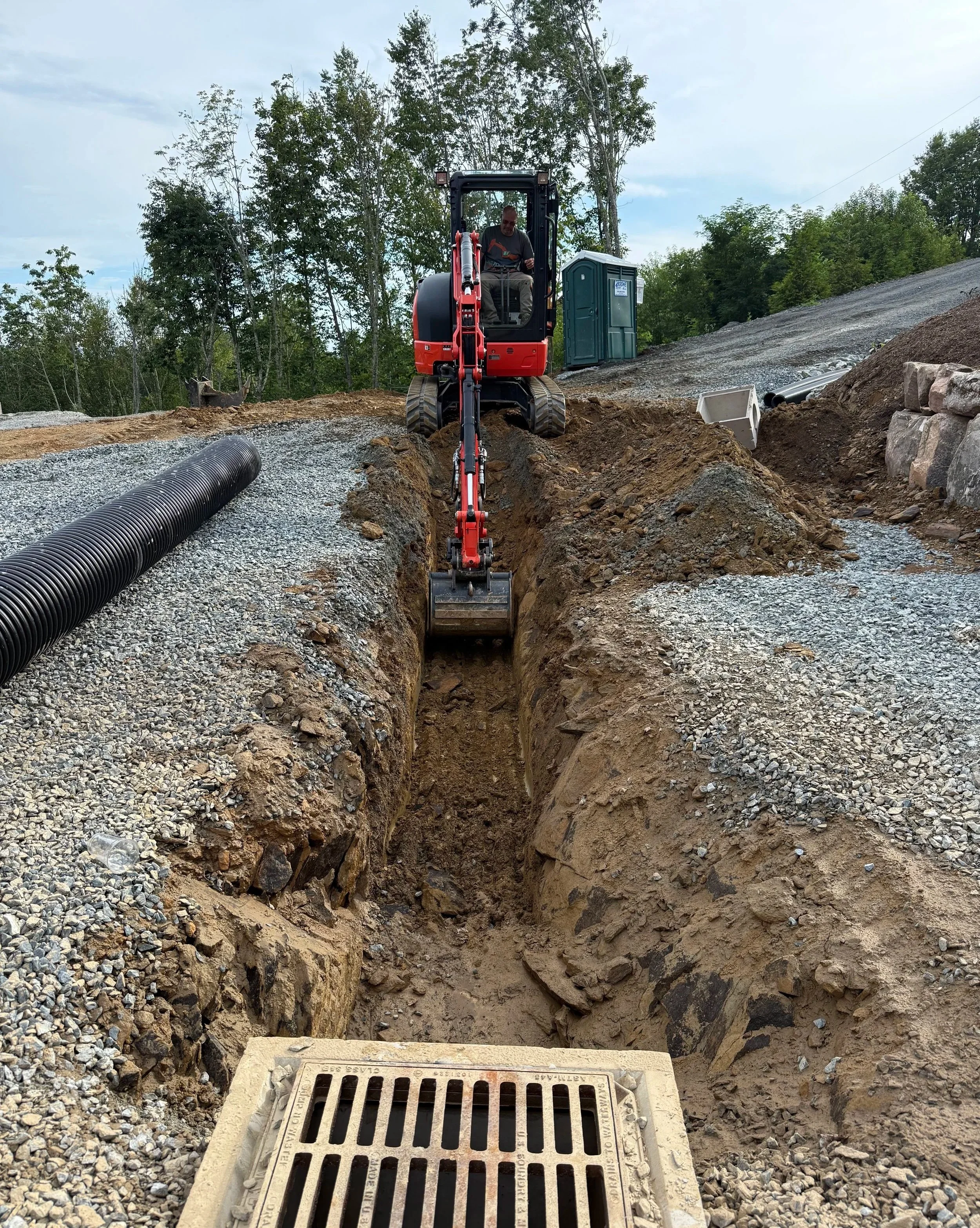 A construction worker operating a small excavator on a dirt and gravel surface near a portable toilet, with trees in the background and a stormy sky overhead.