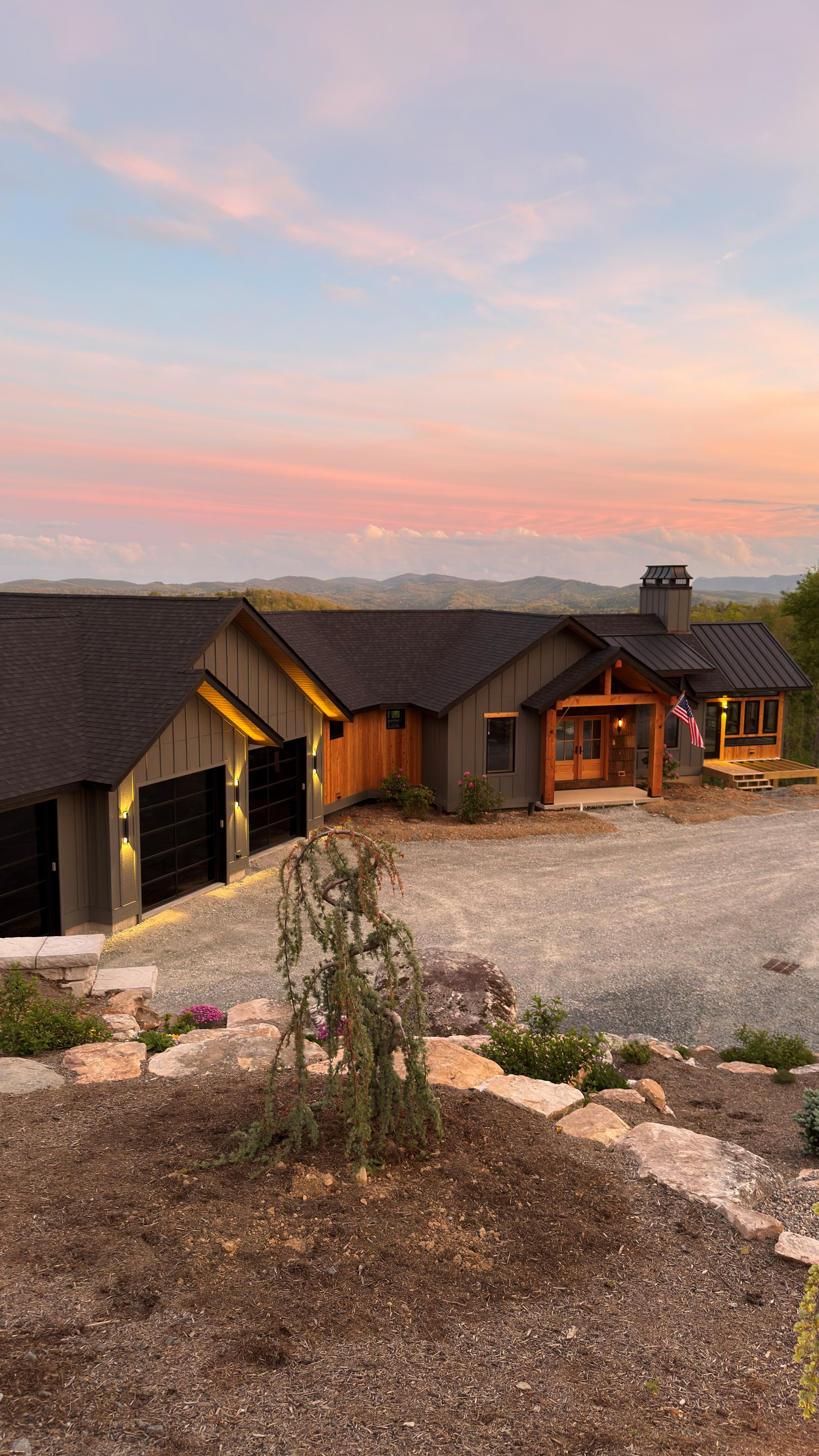 A modern house with a black roof, wooden accents, and a front porch with an American flag. The house is surrounded by a gravel driveway, landscaped yard with rocks, plants, and flowers. Background shows rolling hills and a colorful sky at sunset.