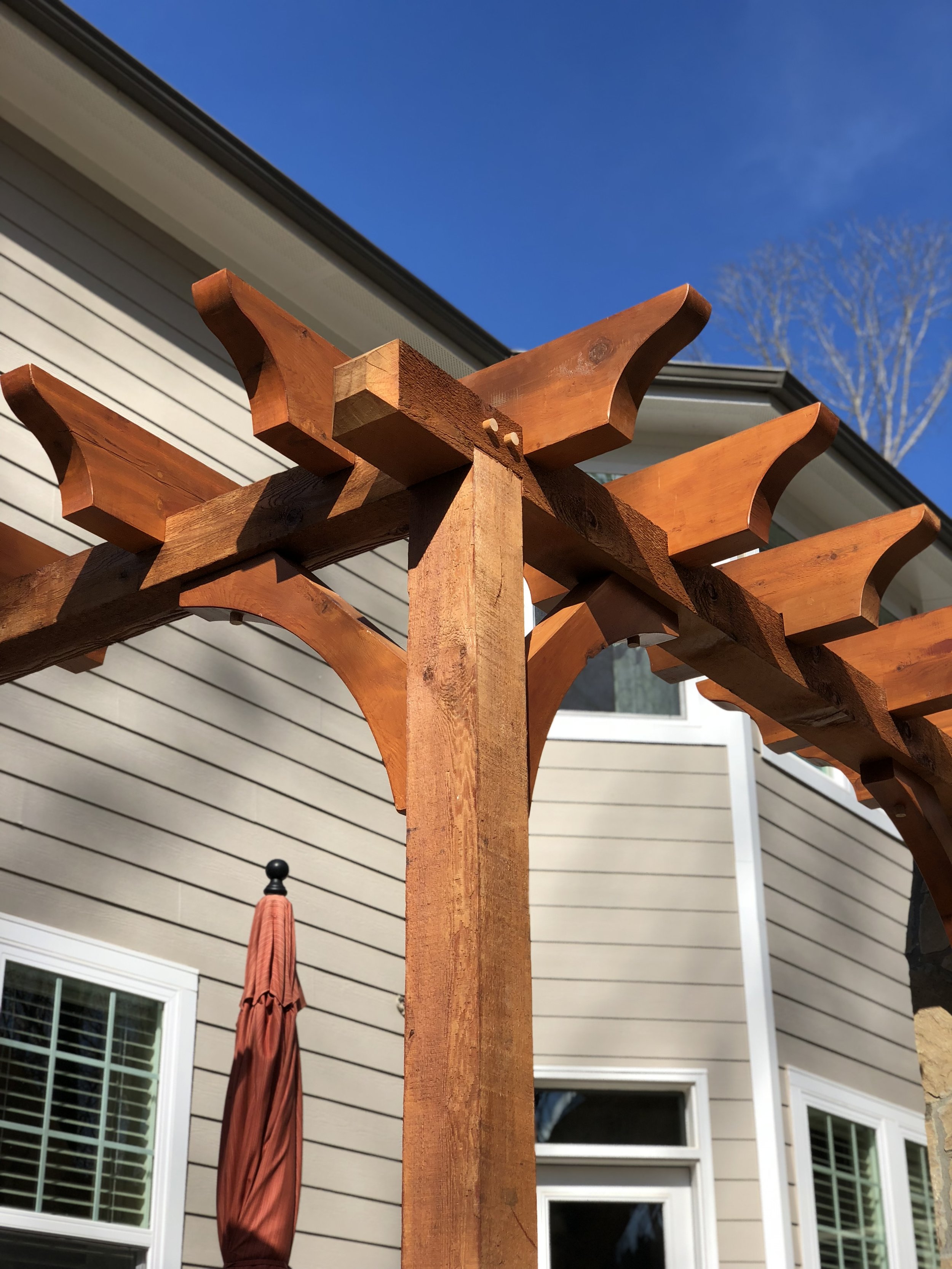 Wooden pergola structure with a closed red patio umbrella hanging underneath, set in a backyard with beige siding and white window frames, under a clear blue sky.