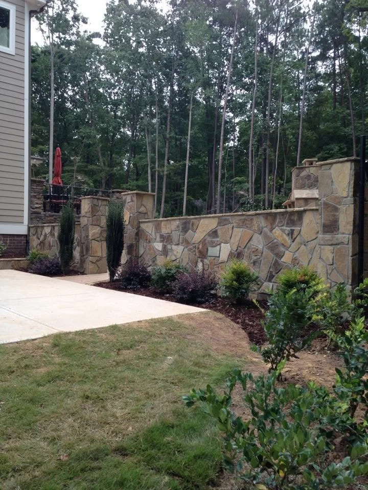 A backyard with a stone retaining wall, small plants and bushes, a concrete patio, and a wooded area in the background.