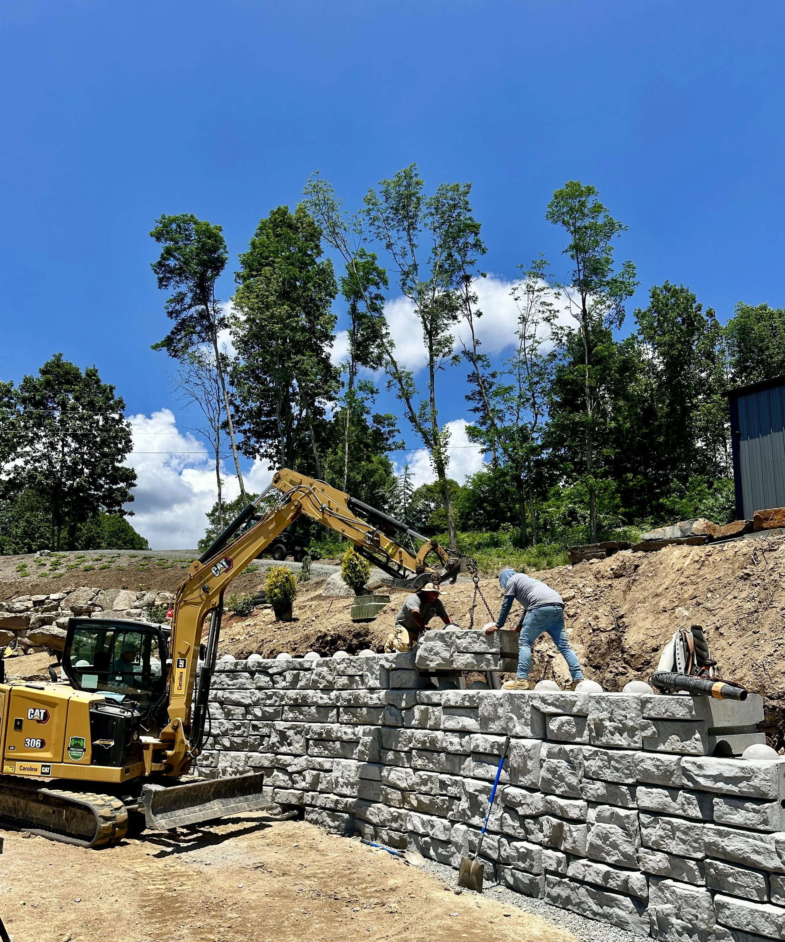 Concrete block retaining wall being constructed with workers, a yellow Caterpillar excavator, and a backdrop of trees and clear blue sky.