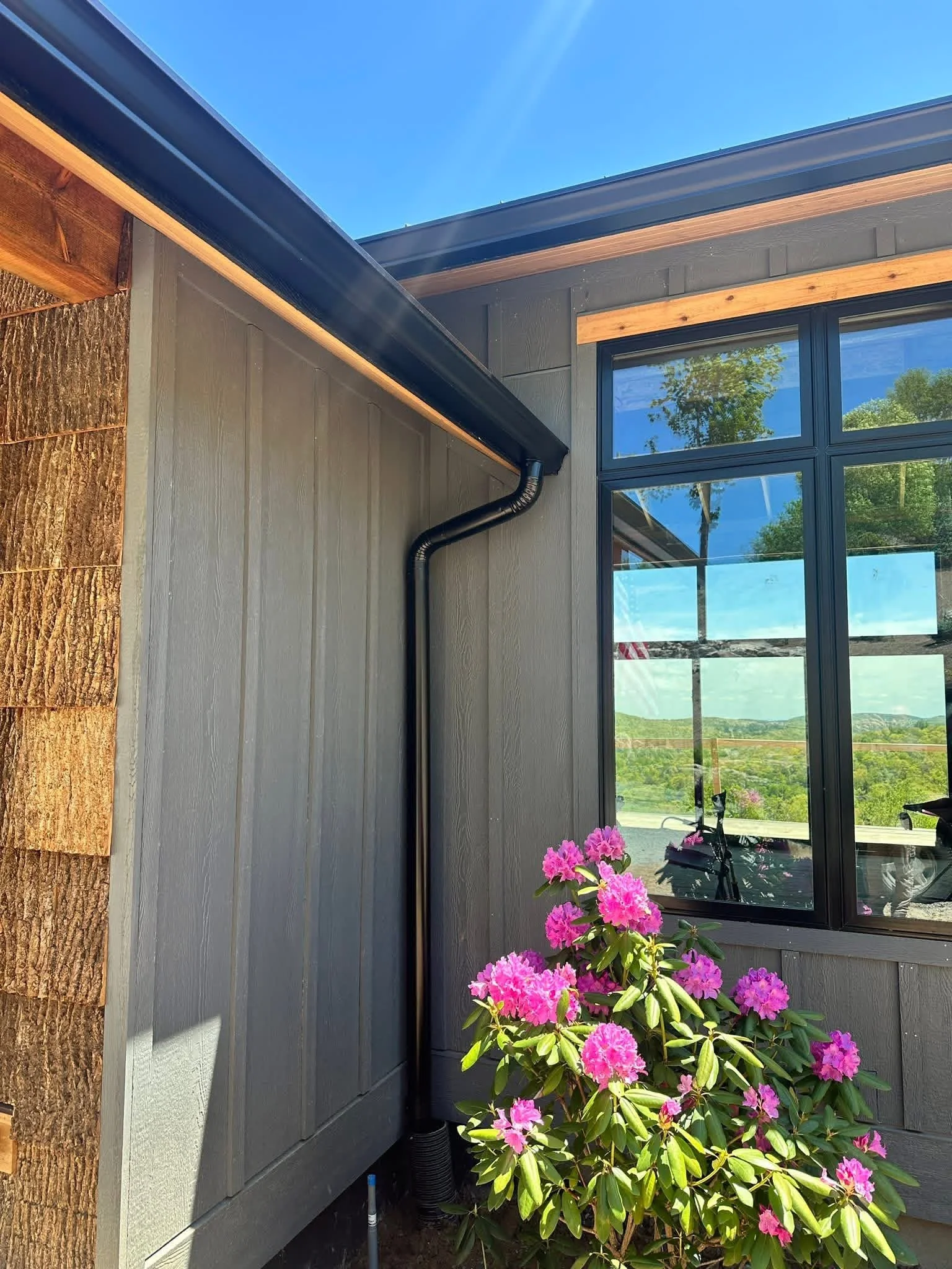 A corner of a house showing a window with a view of trees and hills, a pink flowering plant in front, gray and wooden exterior walls, a black gutter, and clear blue sky.