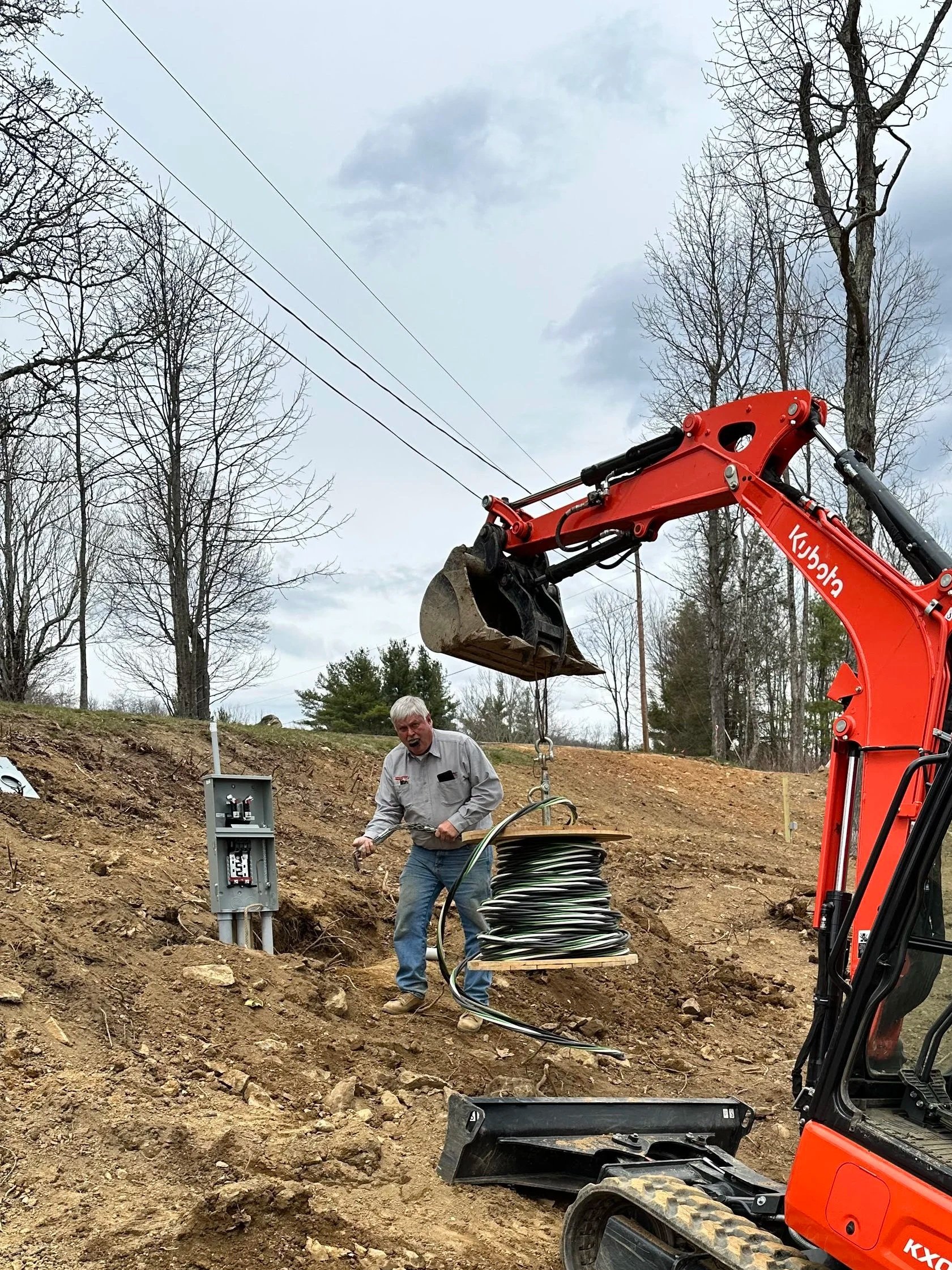 A construction worker stands outdoors in a dirt area with trees and cloudy sky, holding a coil of electrical cable. An orange Kubota excavator is lifting a bucket with a wire or cable above the worker, near an electrical box on a wood post.
