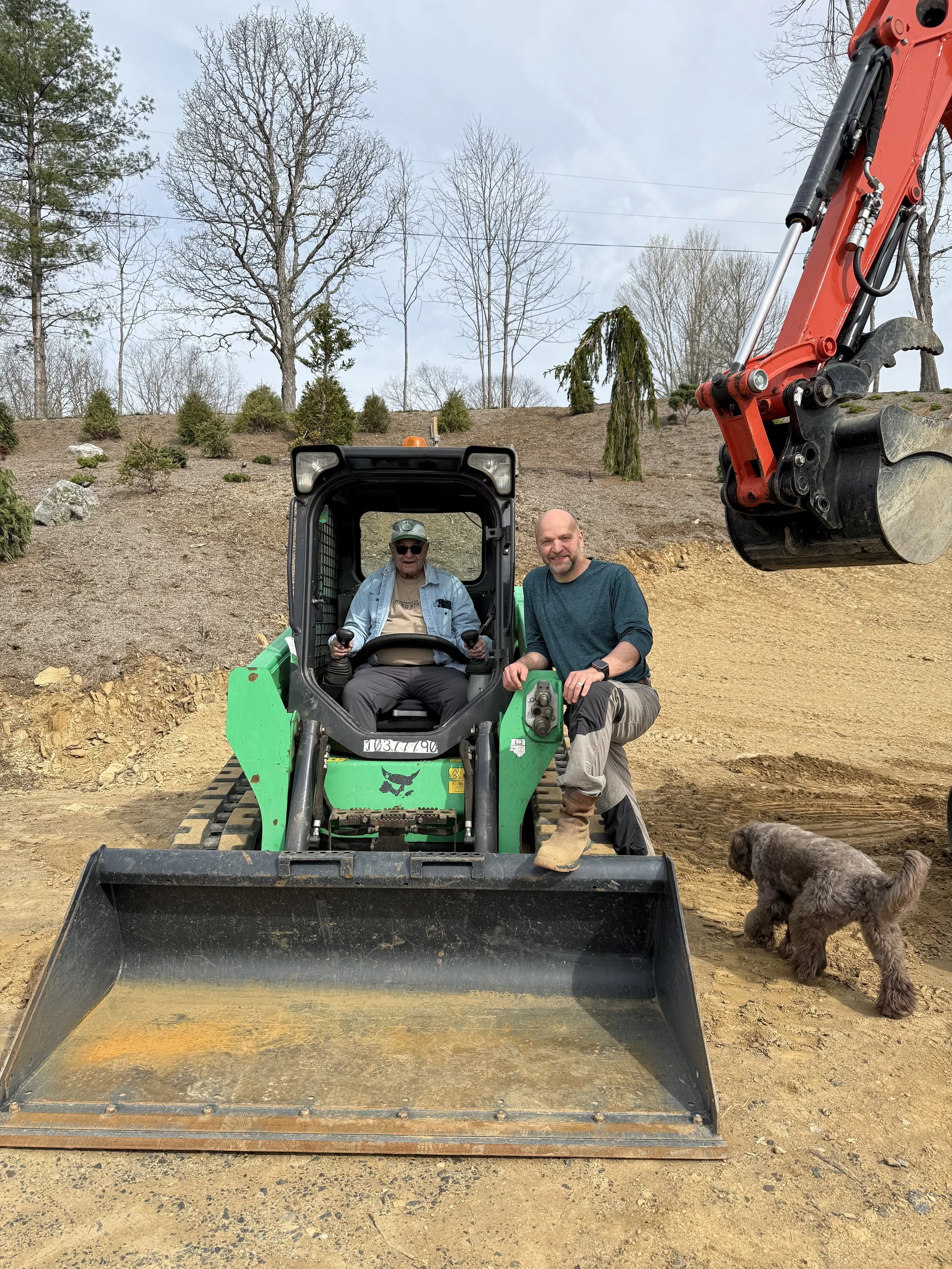 Two men, one sitting in a small green and black excavator and the other standing beside it, in a dirt area with trees and shrubs in the background. A dog is on the ground near the excavator, and an orange construction vehicle with a hydraulic arm is visible in the upper right corner.