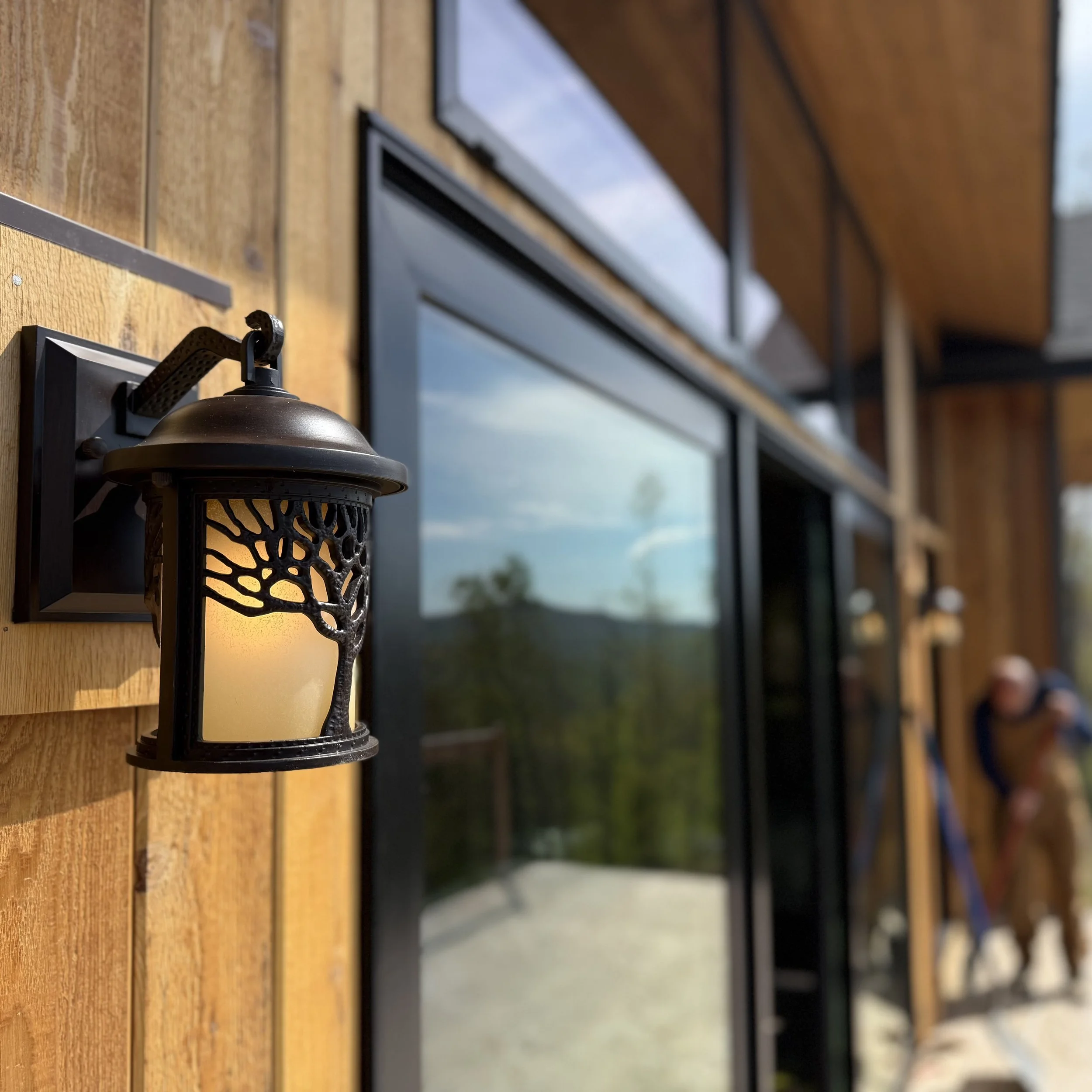Close-up of a black outdoor wall lantern attached to a wooden exterior wall with black trim, against a backdrop of a building with large windows and an outdoor landscape.