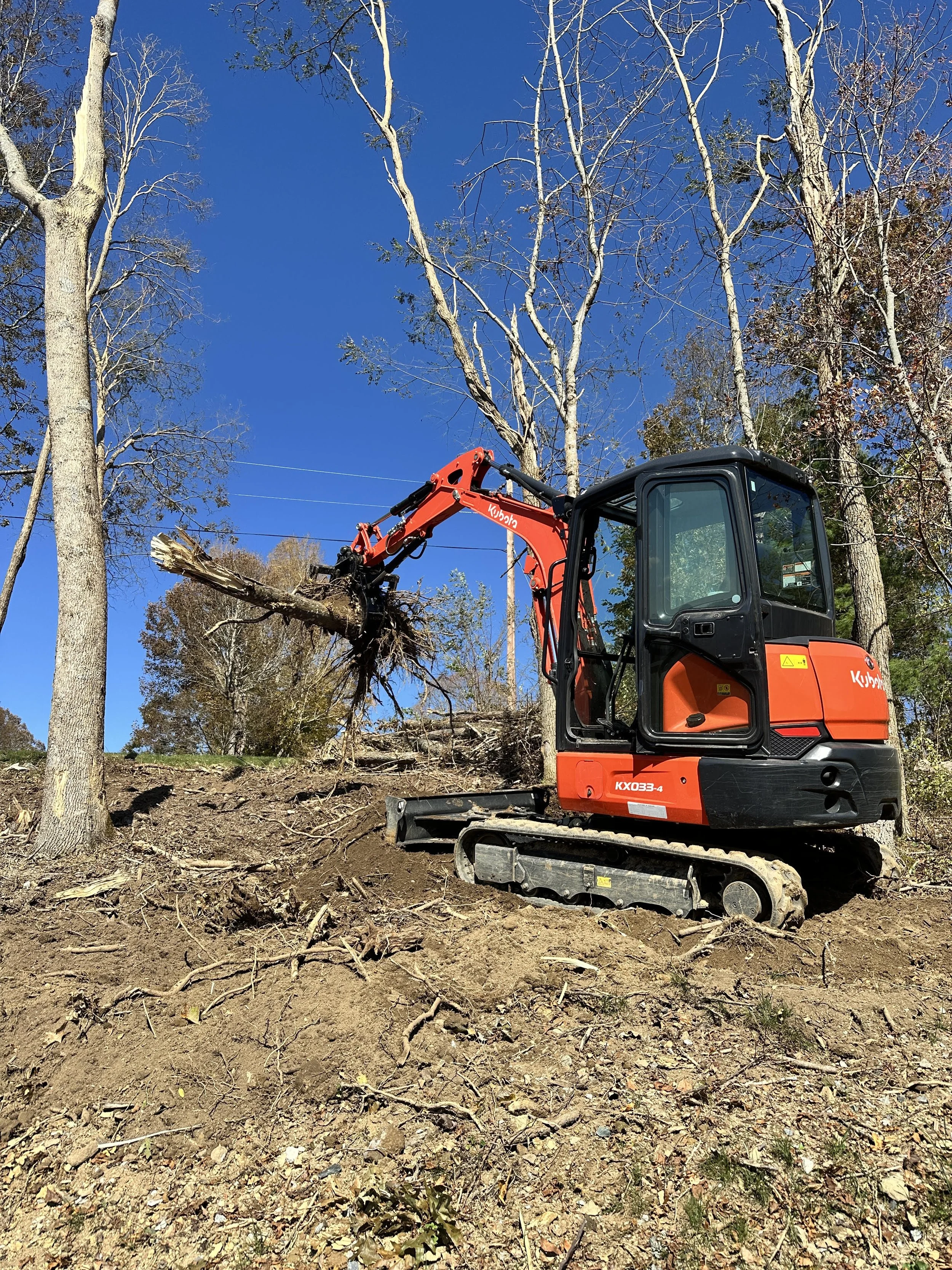 A small orange bulldozer or excavator with tracks, clearing land with trees around, under a clear blue sky.