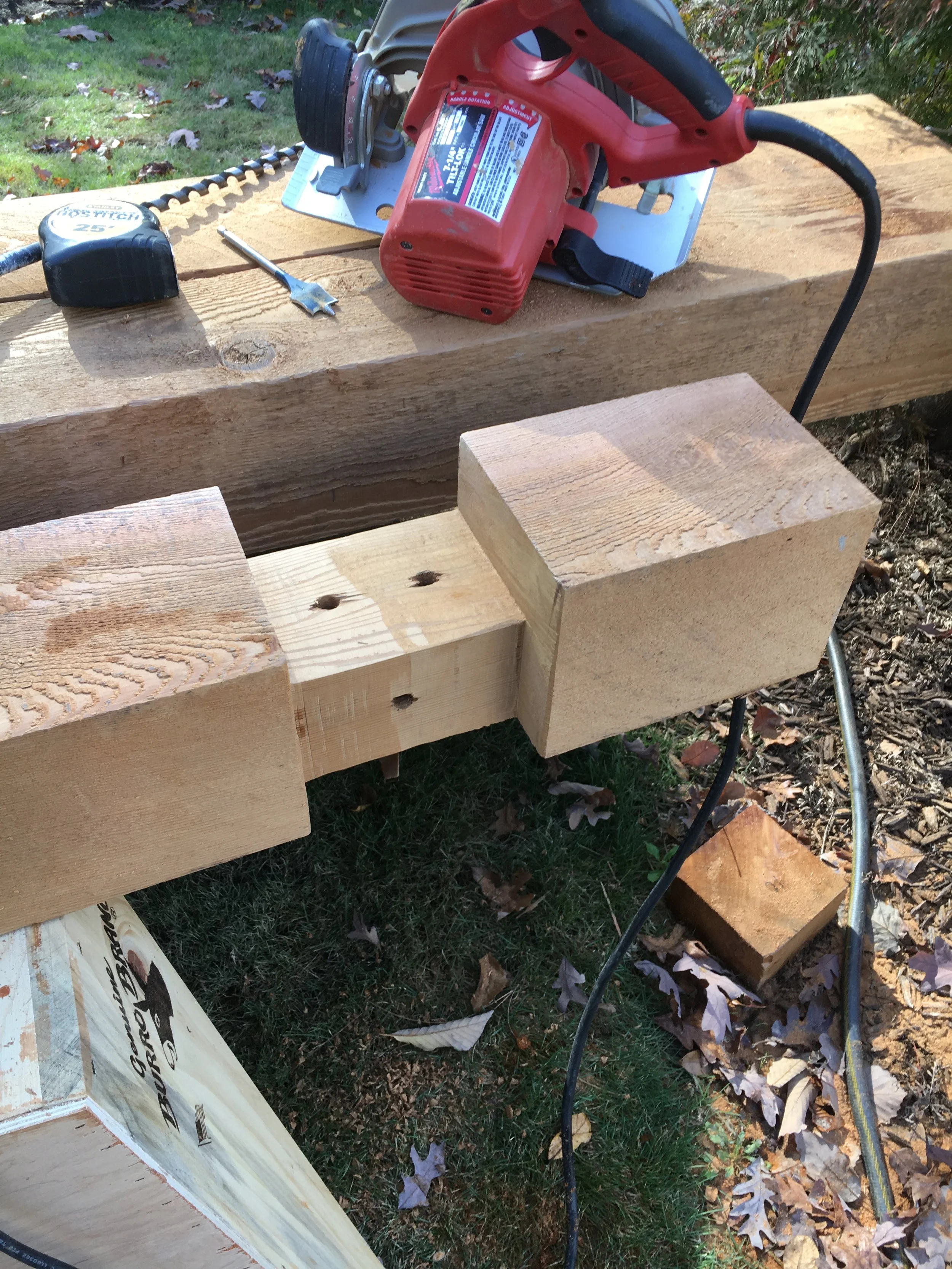 A red cordless power drill, a tape measure, and a flat-head screwdriver on a wooden workbench outdoors. The workbench is supported by sawhorses, and there are wooden blocks attached to the sides. Fallen leaves are on the grass nearby.