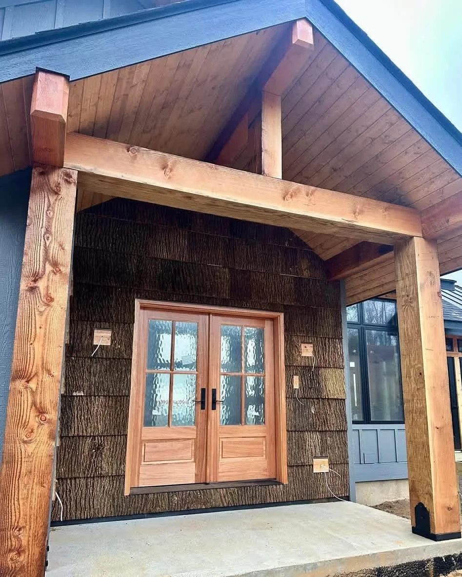 Front porch of a house with a wood frame, dark brown wooden siding, and double glass-paneled wooden doors. The porch has a concrete floor and an overhanging roof with wood paneling.