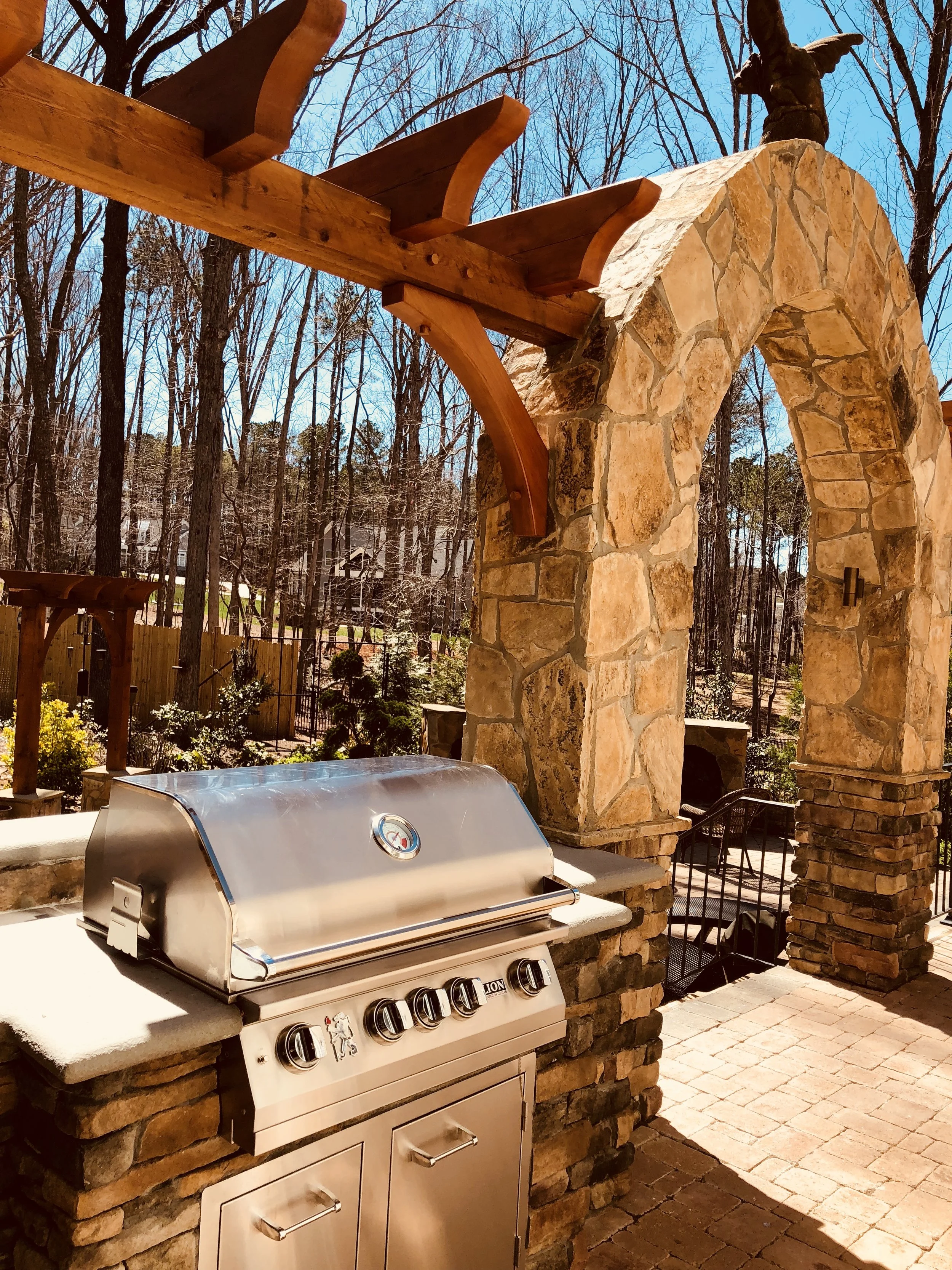 A stainless steel outdoor grill on a kitchen counter with stone pillars and a stone archway in a backyard. Trees and a wooden fence are in the background, and a blue sky is visible.