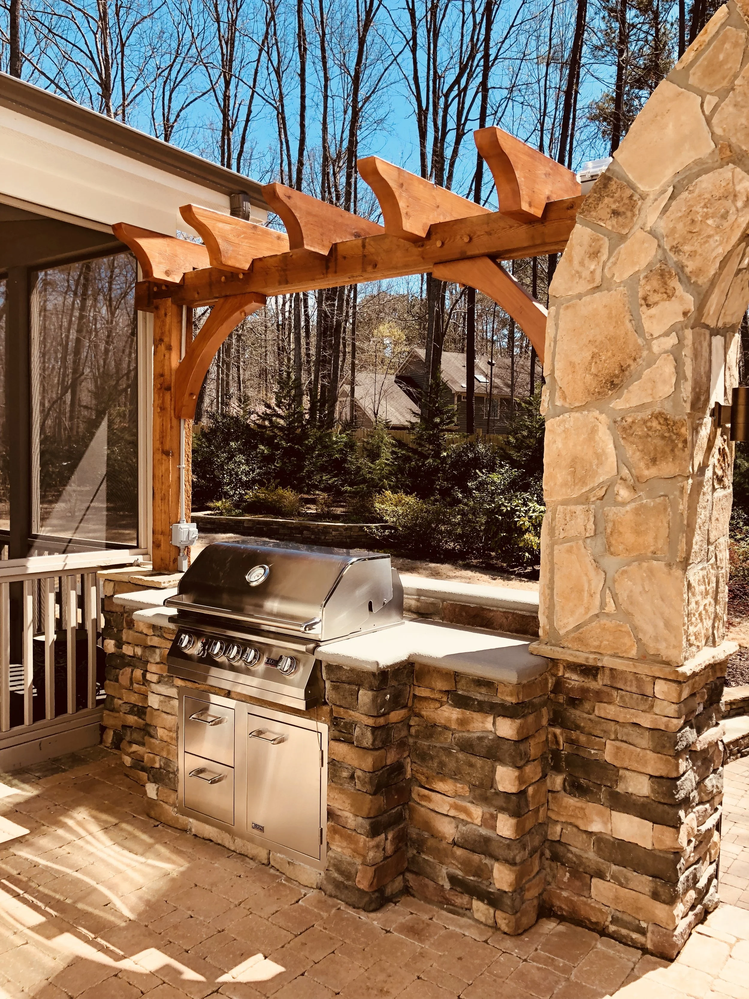 Outdoor kitchen with a stainless steel grill built into a stone countertop, adjacent to a stone pillar, all under a wooden pergola with curved supports, in a backyard with trees and neighboring houses in the background.