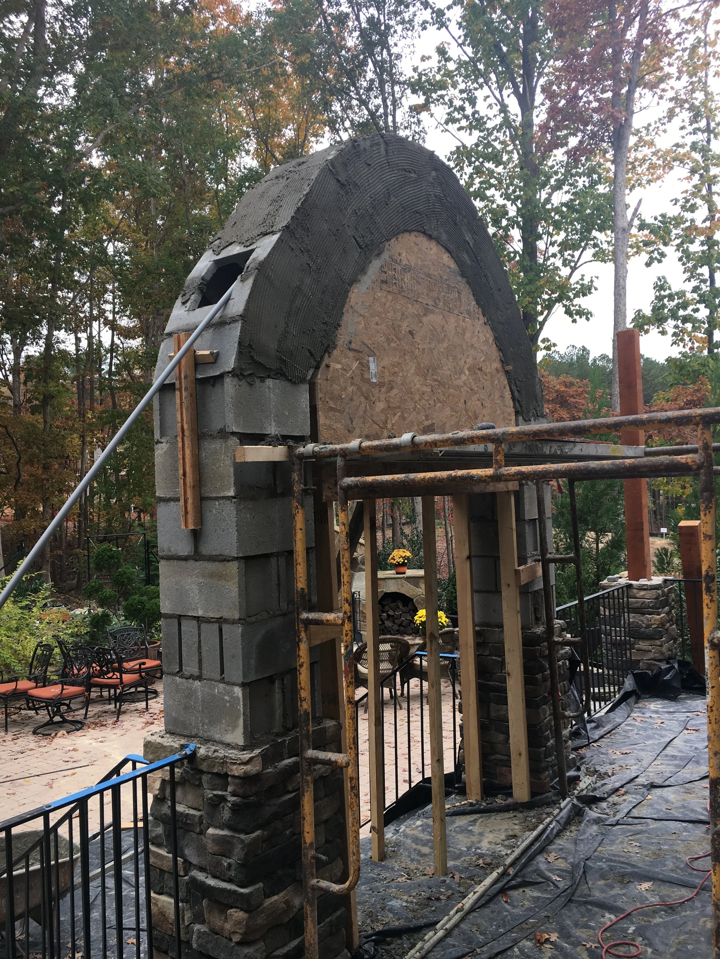 Construction site of a stone archway with scaffolding and building materials, surrounded by trees with fall foliage.