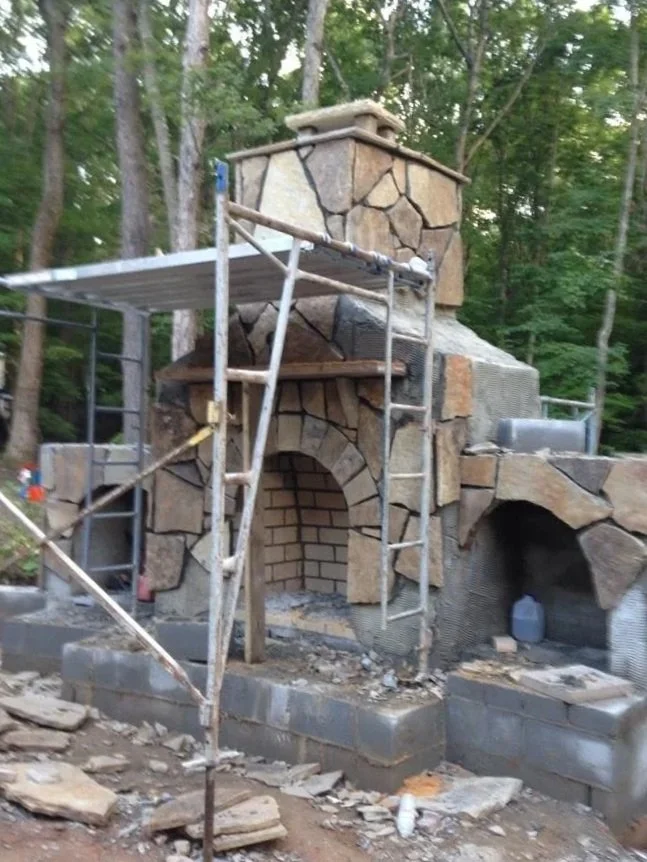 Construction site of a stone and brick outdoor fireplace with scaffolding around it, surrounded by trees.