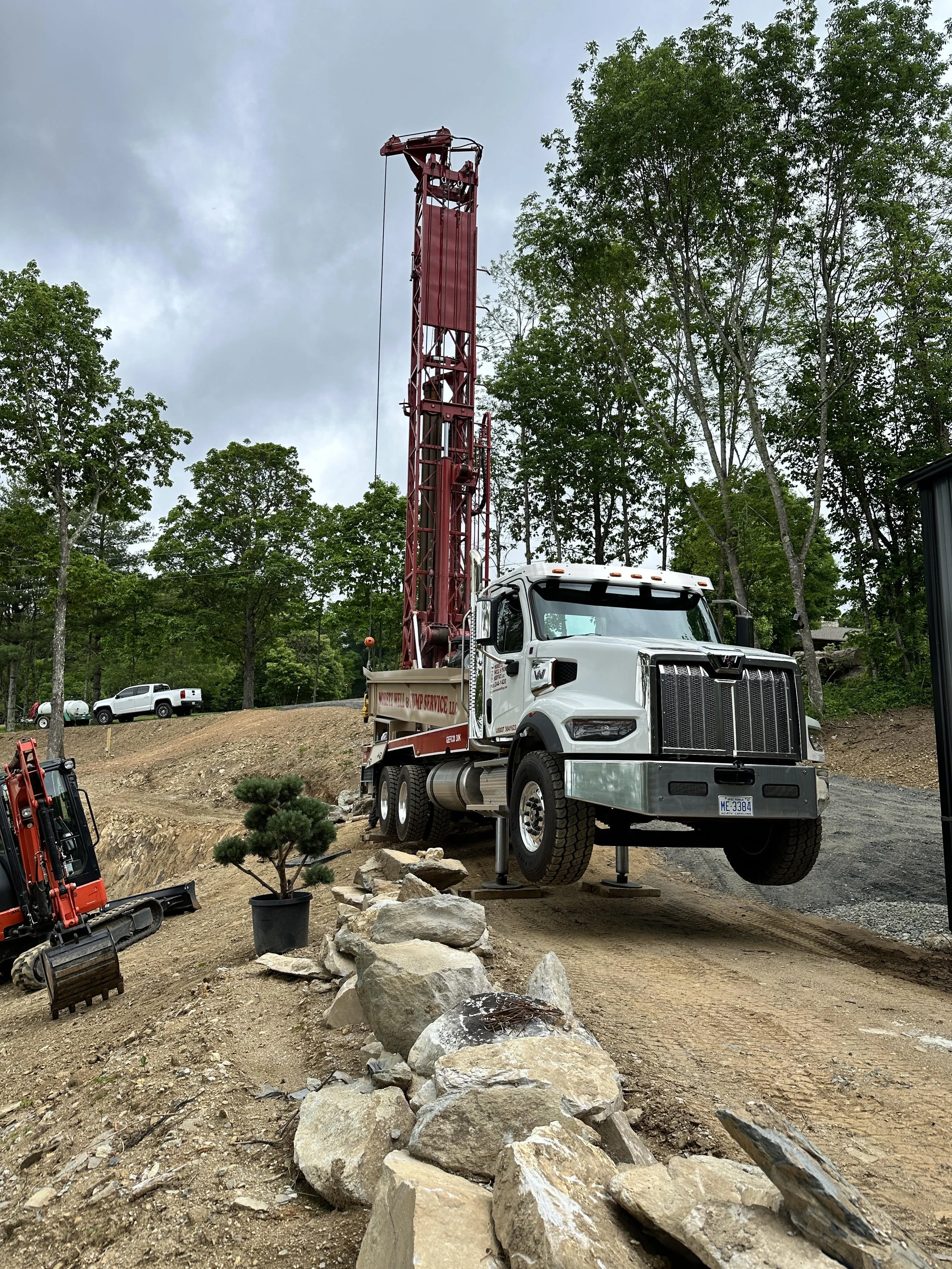 A construction site with a large red drilling truck lifted off the ground, supported by metal stands, surrounded by rocks, a small potted tree, and other construction equipment, with a backdrop of green trees and cloudy sky.