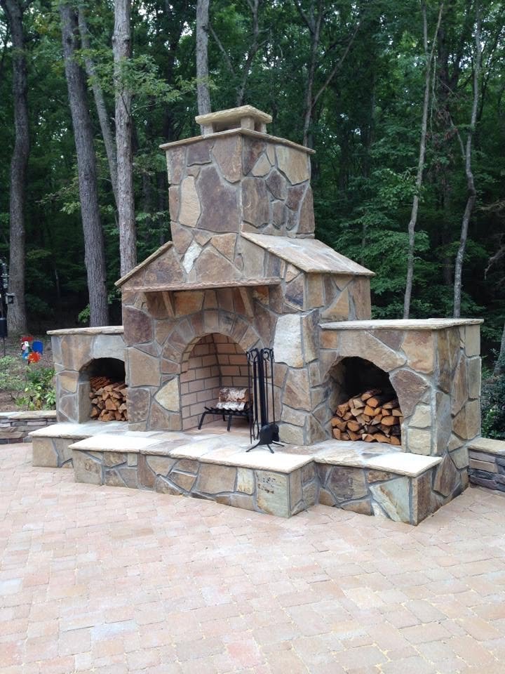 Stone outdoor fireplace with wood storage compartments on each side, situated on a brick patio in a wooded backyard.