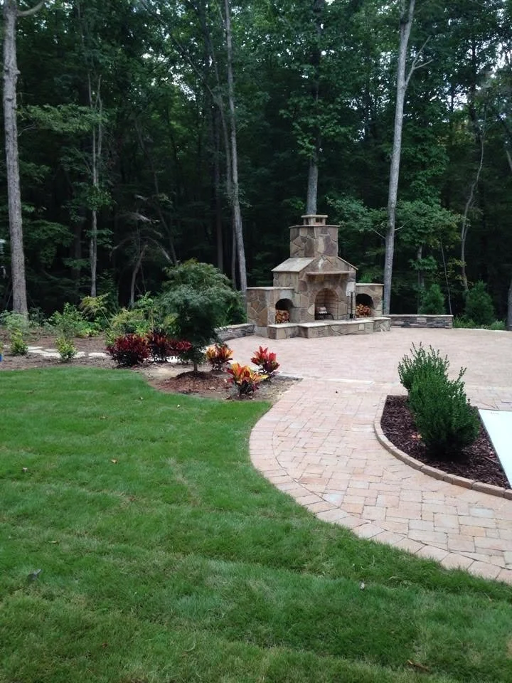 A backyard with a curved brick patio, a stone outdoor fireplace, and landscaped garden beds with bushes and plants, surrounded by tall trees.