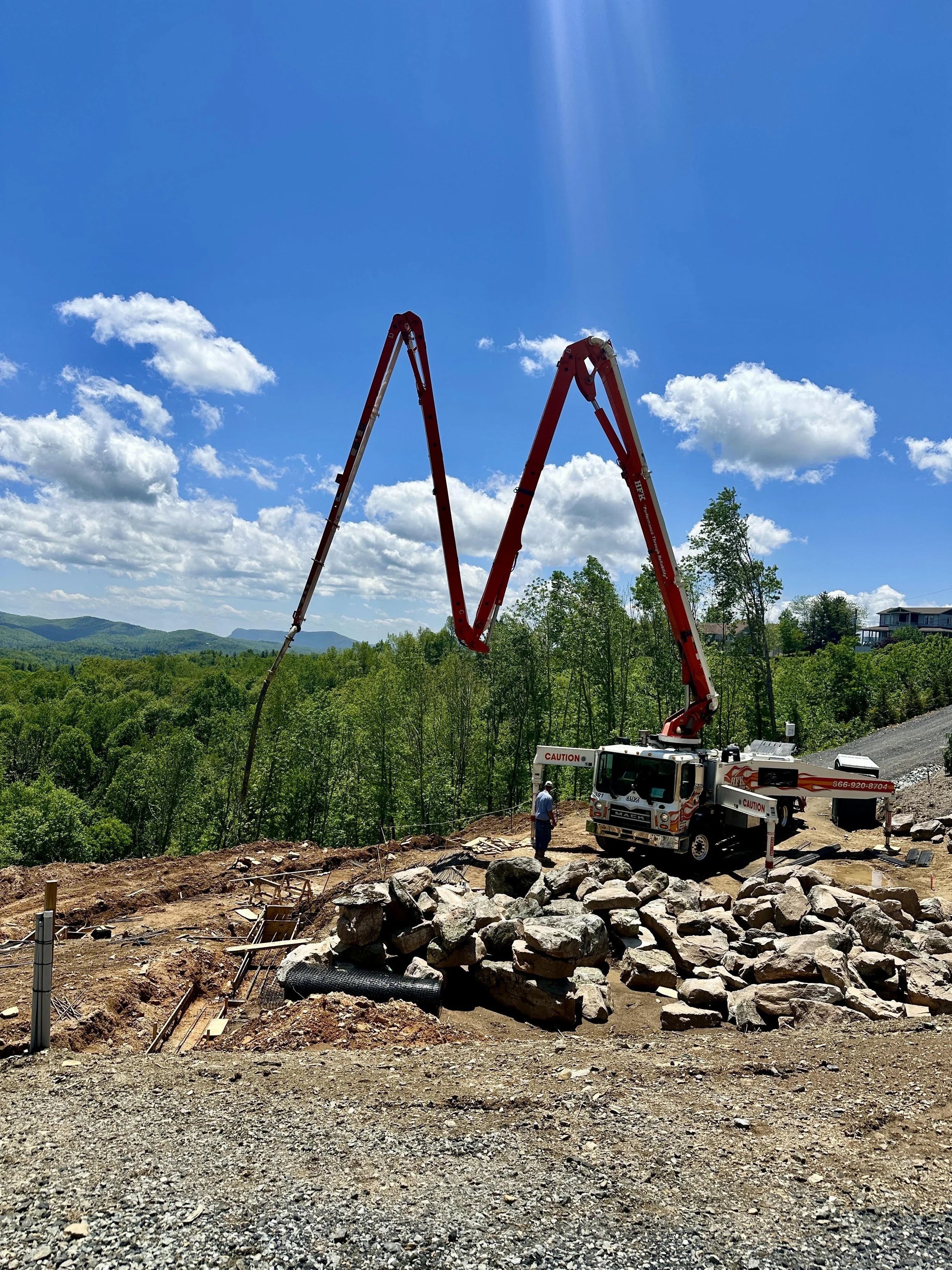Construction site with a large red concrete pump truck pouring concrete, rocks, gravel, and construction materials on uneven ground with green trees and hills in the background under a blue sky with some clouds.