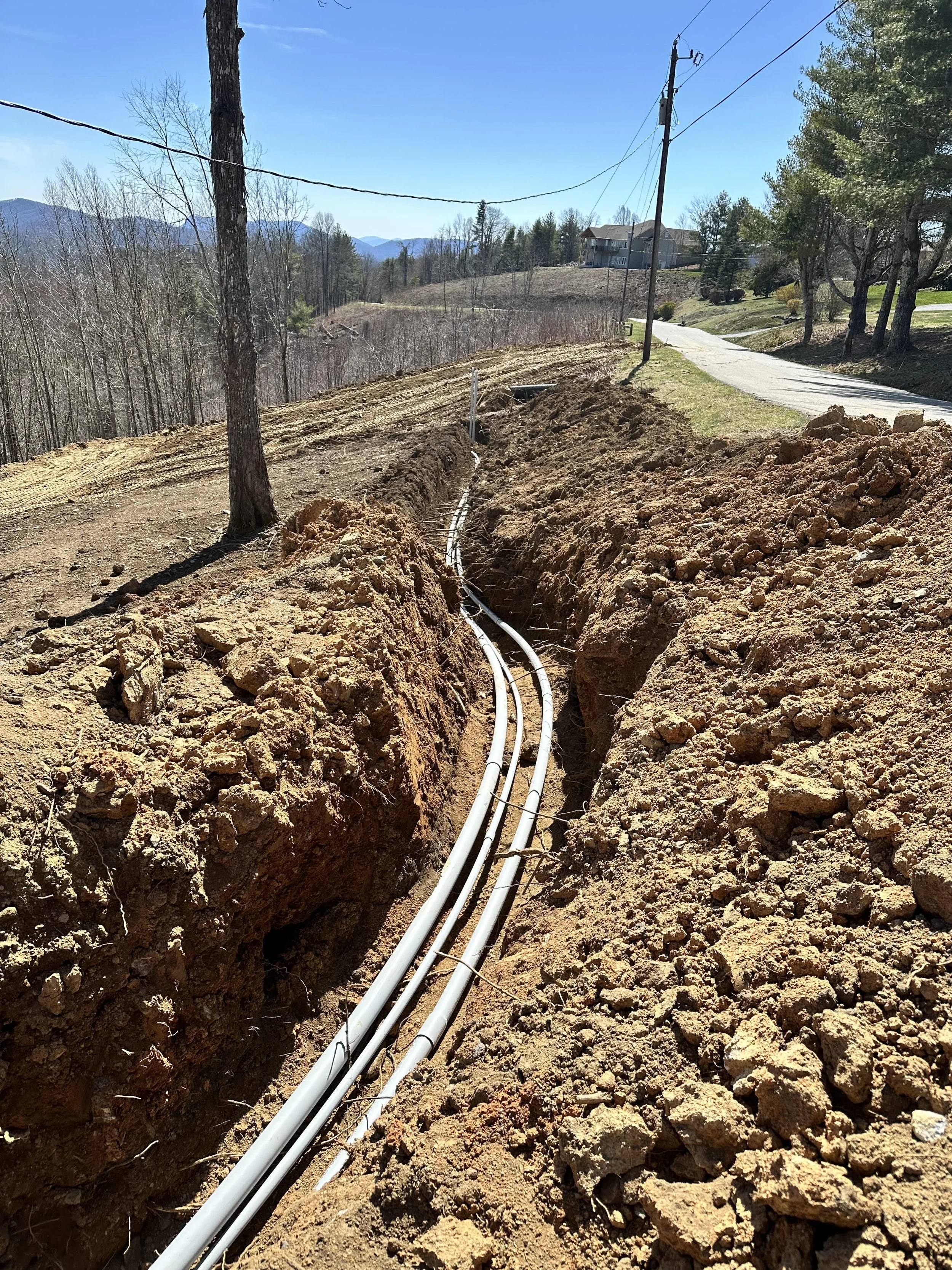 Construction site with white pipes installed in a trench, dirt piles on the sides, trees, a paved road, and houses in a mountainous background under a clear blue sky.