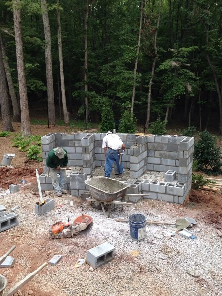 Two workers building a cinder block wall outdoors in a wooded area, with construction tools and materials scattered around.