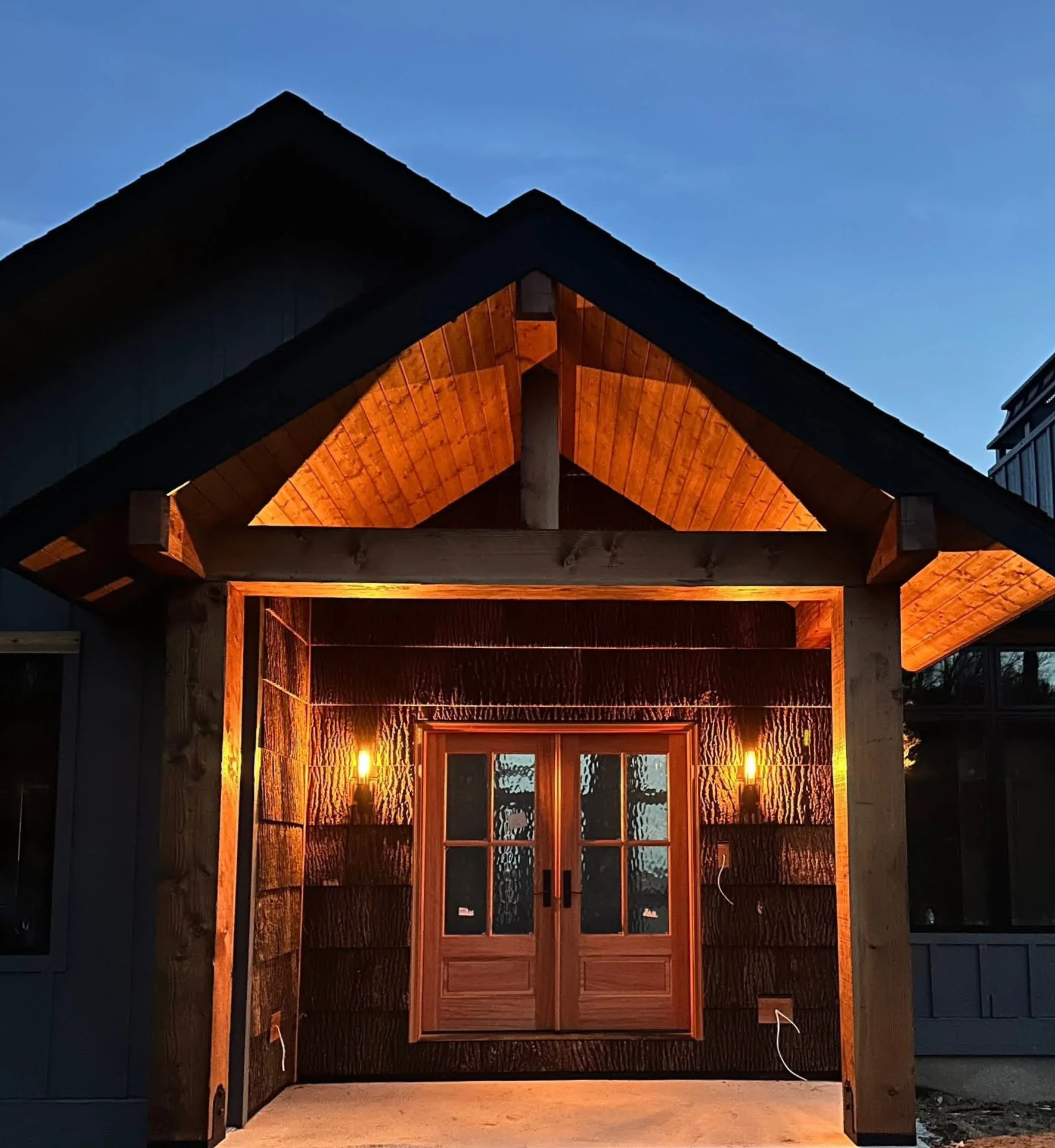 View of a partially constructed or renovated outdoor cabin or shed with a peaked roof. The structure is made of wood and is lit from inside, creating a warm glow through the windows and open framing. The background shows an evening sky.