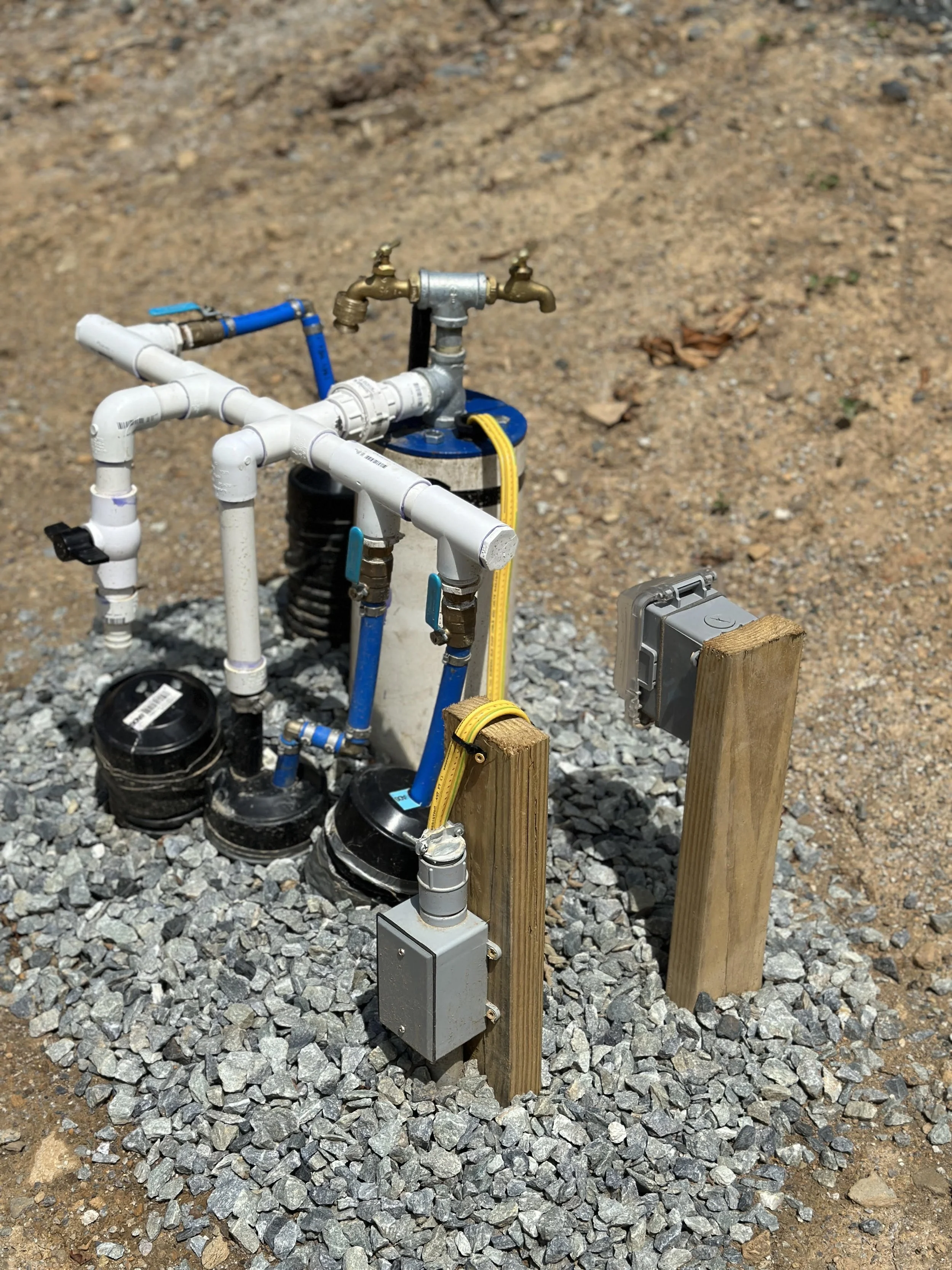 Water meters, pipes, and valves installed outdoors on gravel, with electrical boxes and wooden posts nearby.