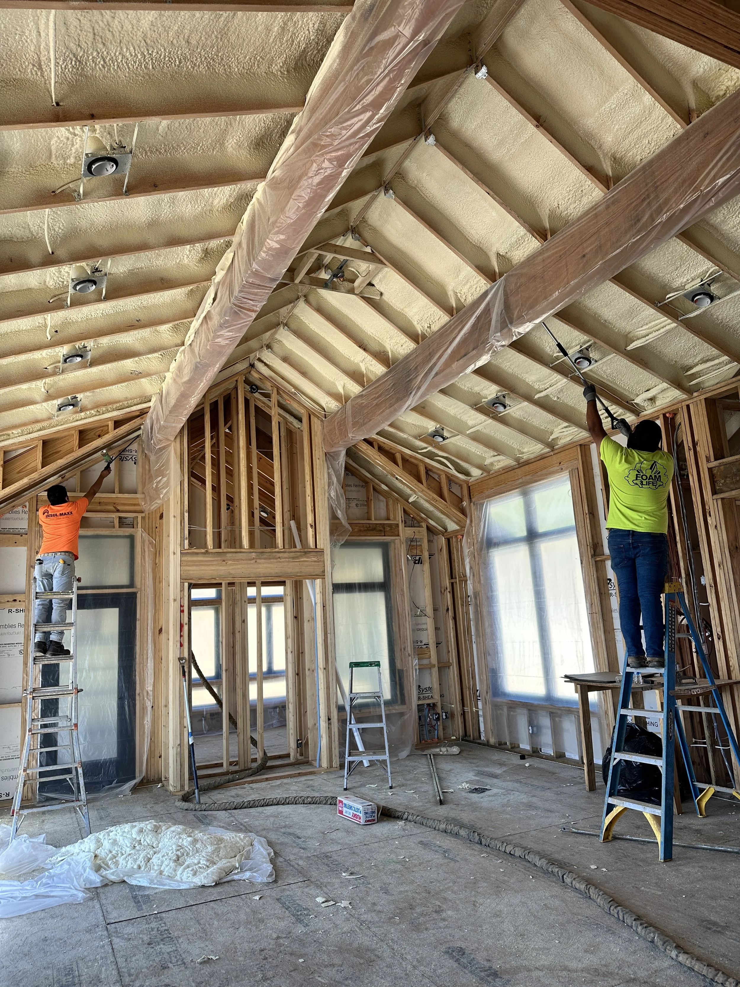 Construction workers installing insulation in the ceiling of a home, using ladders and tools, with wood framing and plastic sheeting around the area.