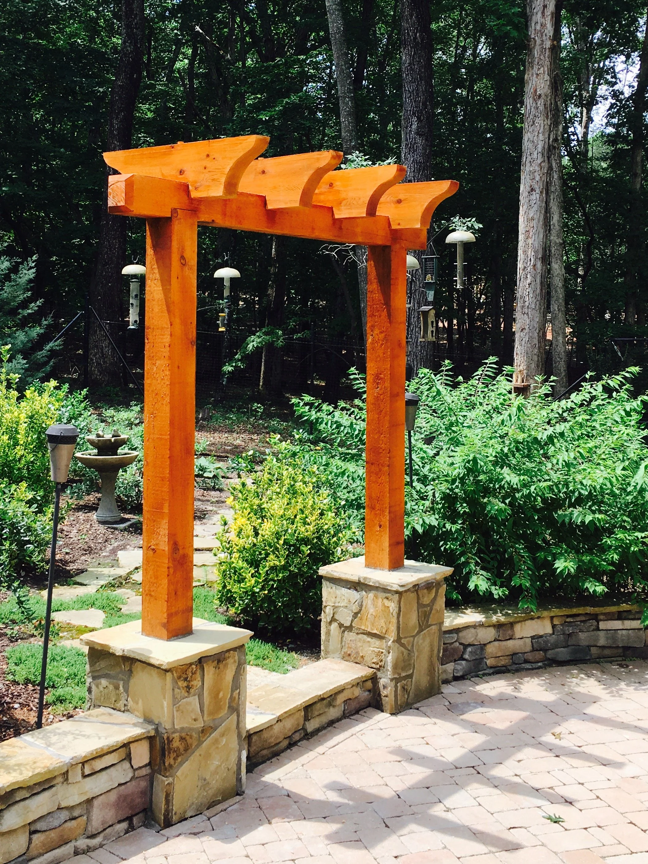 Wooden pergola with stone bases in a garden, surrounded by lush green plants and trees, with a stone pathway.