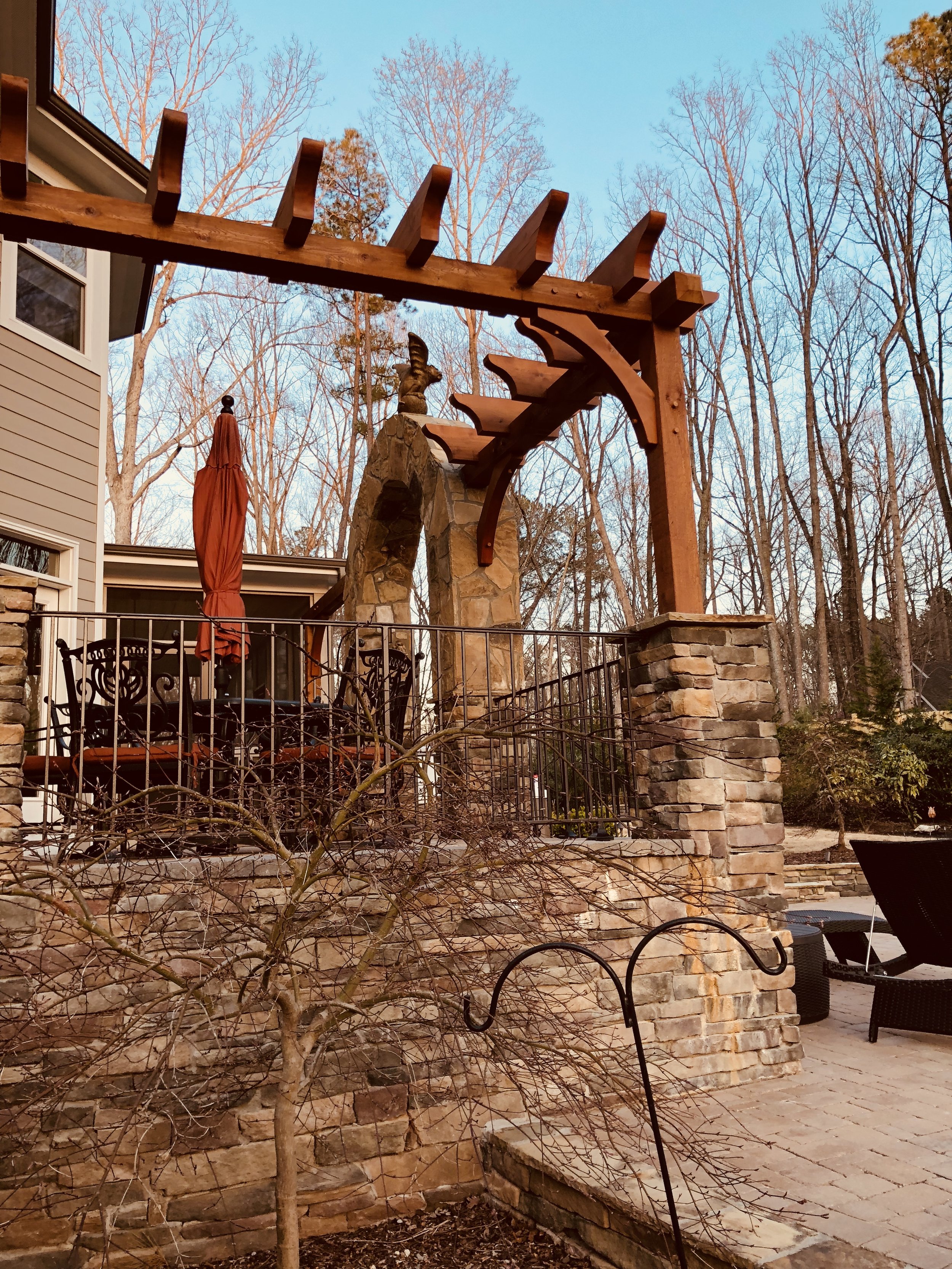 A backyard patio area with a stone wall, metal patio furniture, a leafless tree, and a wooden pergola structure against a backdrop of leafless trees and a blue sky.